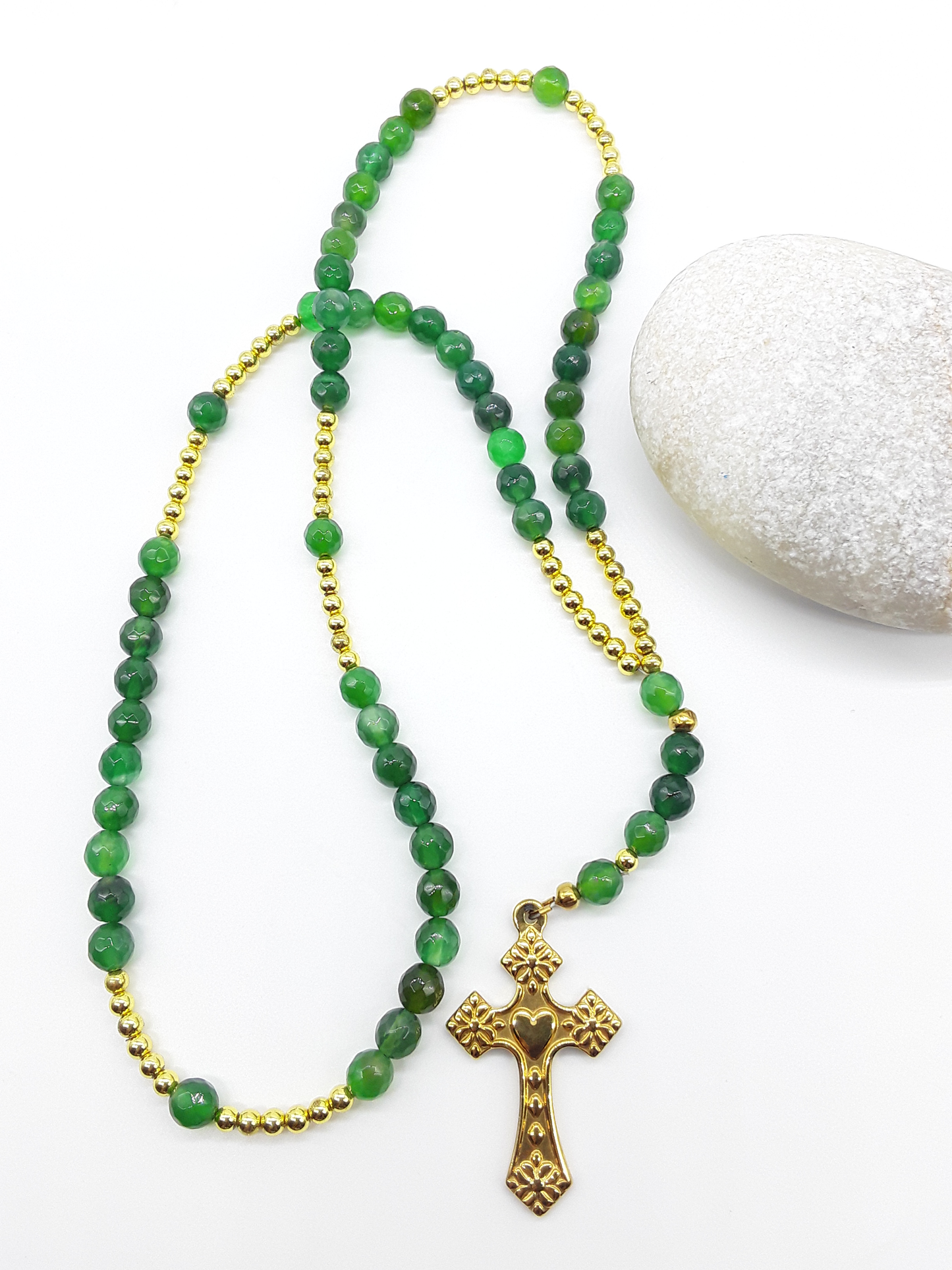 Catholic rosary with natural green agate beads and a stainless steel heart-shaped cross arranged beside a natural stone on a light background.