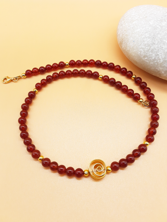 Finding Our Way Carnelian necklace with gold-tone 925 Sterling Silver spiral, beads and clasp by POLMADEVA next to a white stone.
