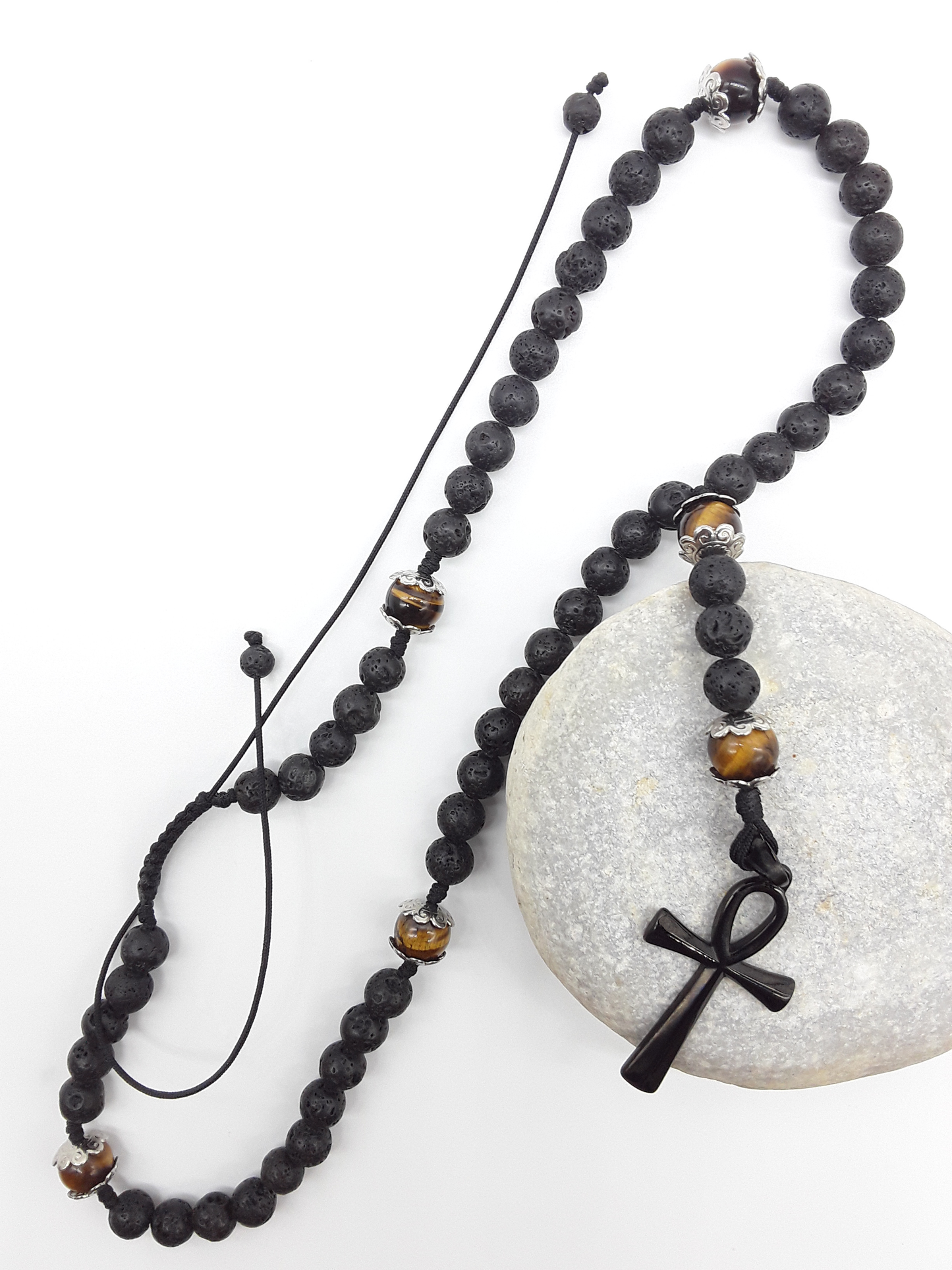 Lava and Tiger’s Eye Catholic Rosary with black stainless steel Ankh cross by Polmadeva resting on natural stone surface.