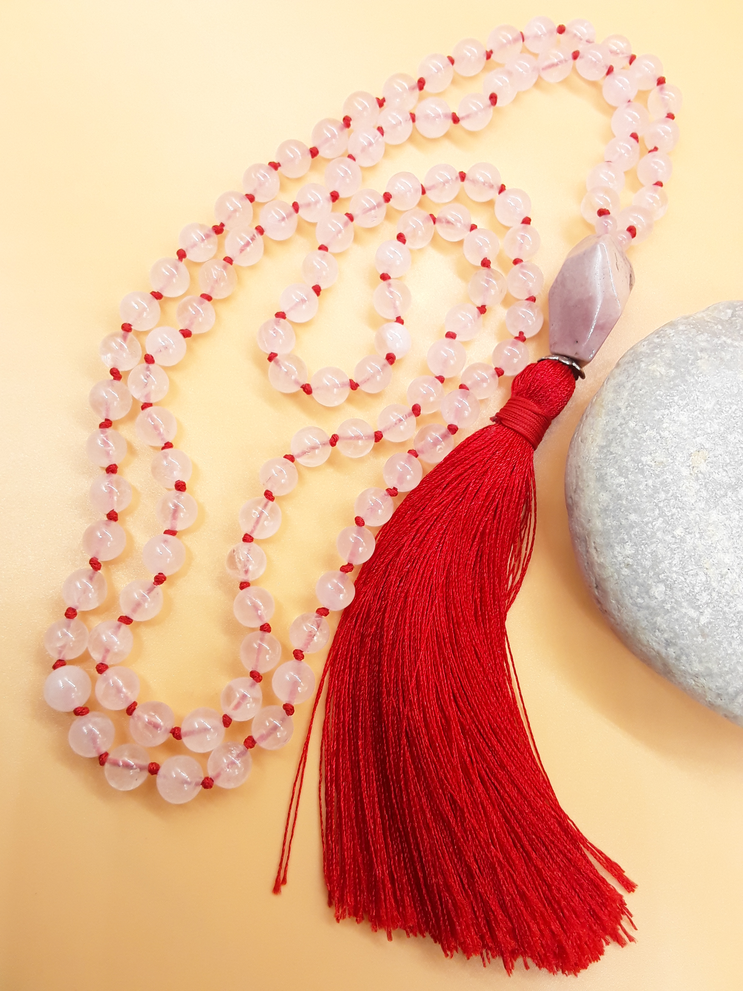 Rose Quartz Mala Beads with Agate guru bead and red tassel placed beside white stone on soft background, Polmadeva