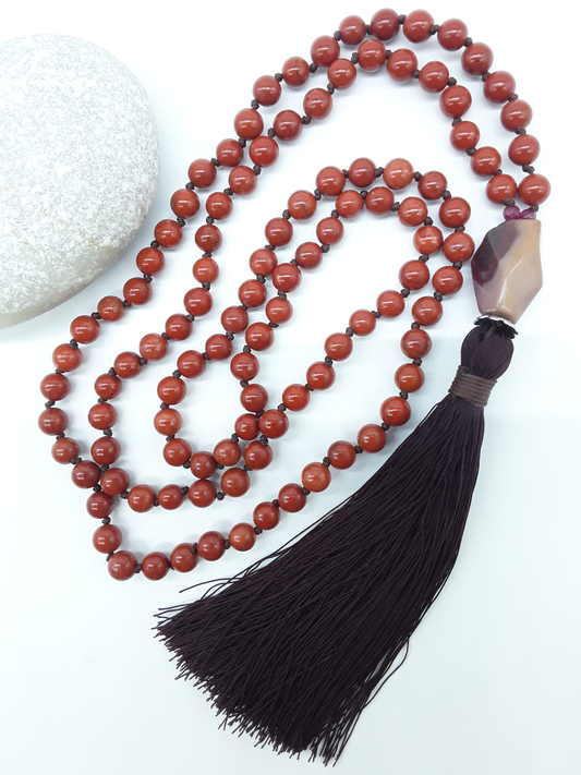 Red Jasper Mala Beads with Agate guru bead and dark tassel placed beside white stone on white background, Polmadeva