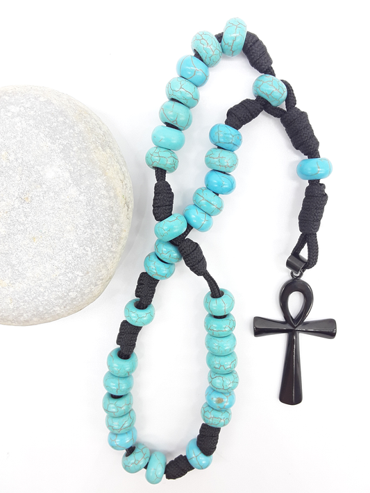 The Living Covenant Paracord Anglican Prayer Beads by Polmadeva beside natural stone, featuring Turquoise and Ankh cross