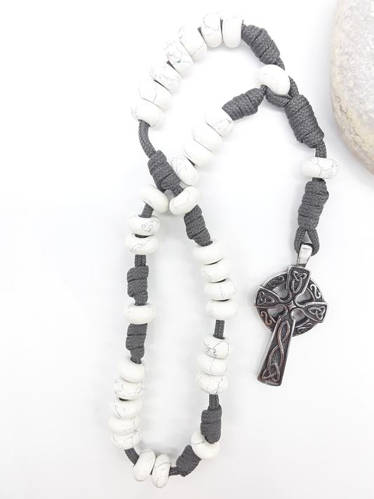 The Quiet Structure Paracord Anglican Prayer Beads by Polmadeva beside natural stone, featuring Howlite and Celtic cross