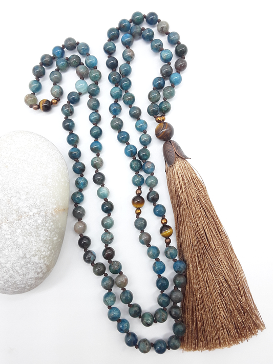 Apatite Mala Beads with Tiger’s Eye, Hematite details and tassel placed beside white stone on white background, Polmadeva