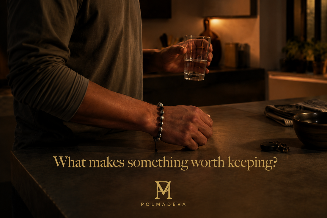 A quiet indoor evening moment where a person stands by a kitchen counter holding a drink, with a bracelet naturally worn on the wrist, warm lighting, and the text “What makes something worth keeping?” alongside the Polmadeva logo.