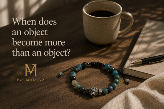 A warm, everyday desk scene with natural light, featuring a Polmadeva bracelet with blue-green beads resting on a wooden table beside a coffee mug and notebook, with the title “When does an object become more than an object?” and the Polmadeva logo.