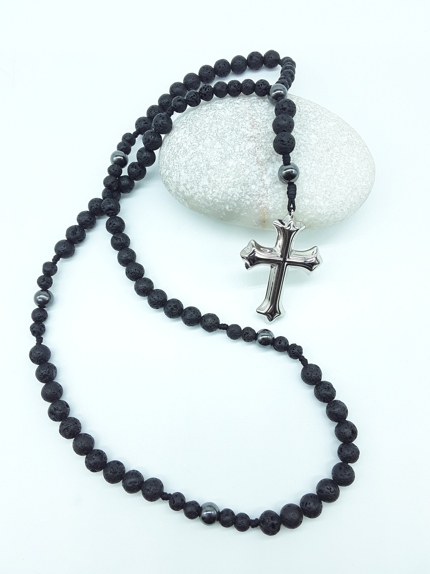 Catholic rosary with lava stone beads, hematite spacers, and a sculpted stainless steel cross shown in full length on a light background over a rock.