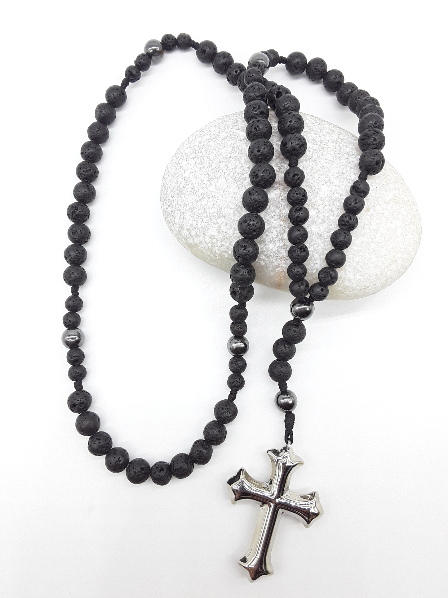 Catholic rosary with lava stone beads, hematite spacers, and a sculpted stainless steel cross shown in full length on a light background over a rock.