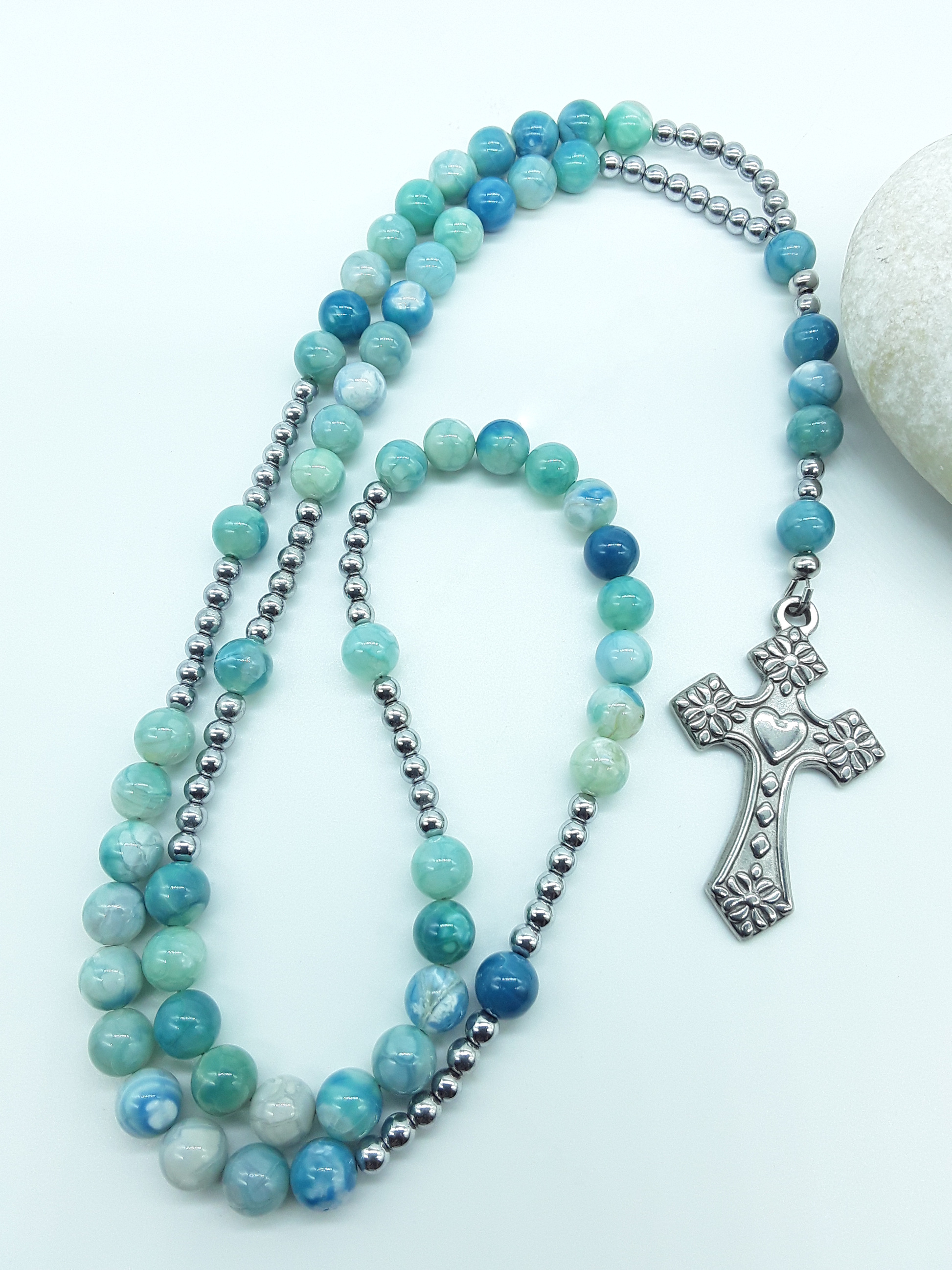 Catholic rosary with natural agate beads and a stainless steel heart-shaped cross arranged beside a natural stone on a light background.
