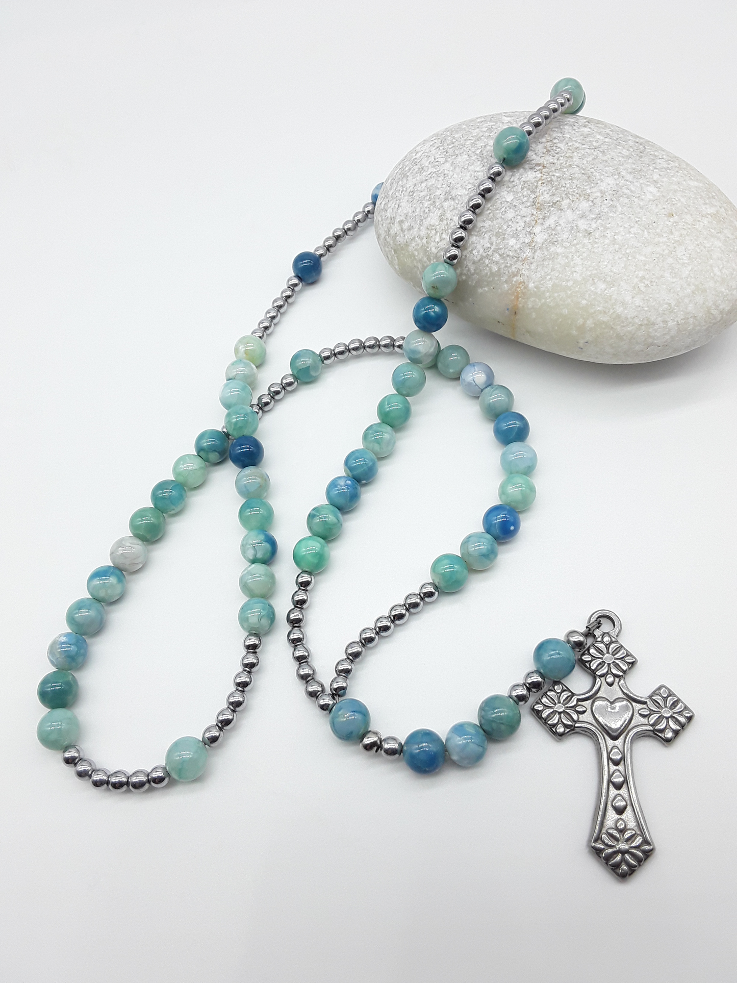 Catholic rosary with natural agate beads and a stainless steel heart-shaped cross arranged beside a natural stone on a light background.
