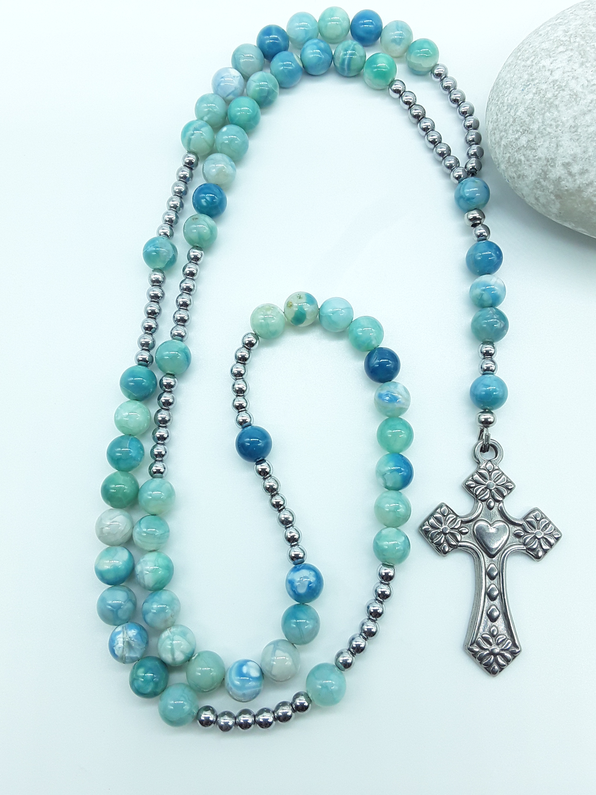 Catholic rosary with natural agate beads and a stainless steel heart-shaped cross arranged beside a natural stone on a light background.
