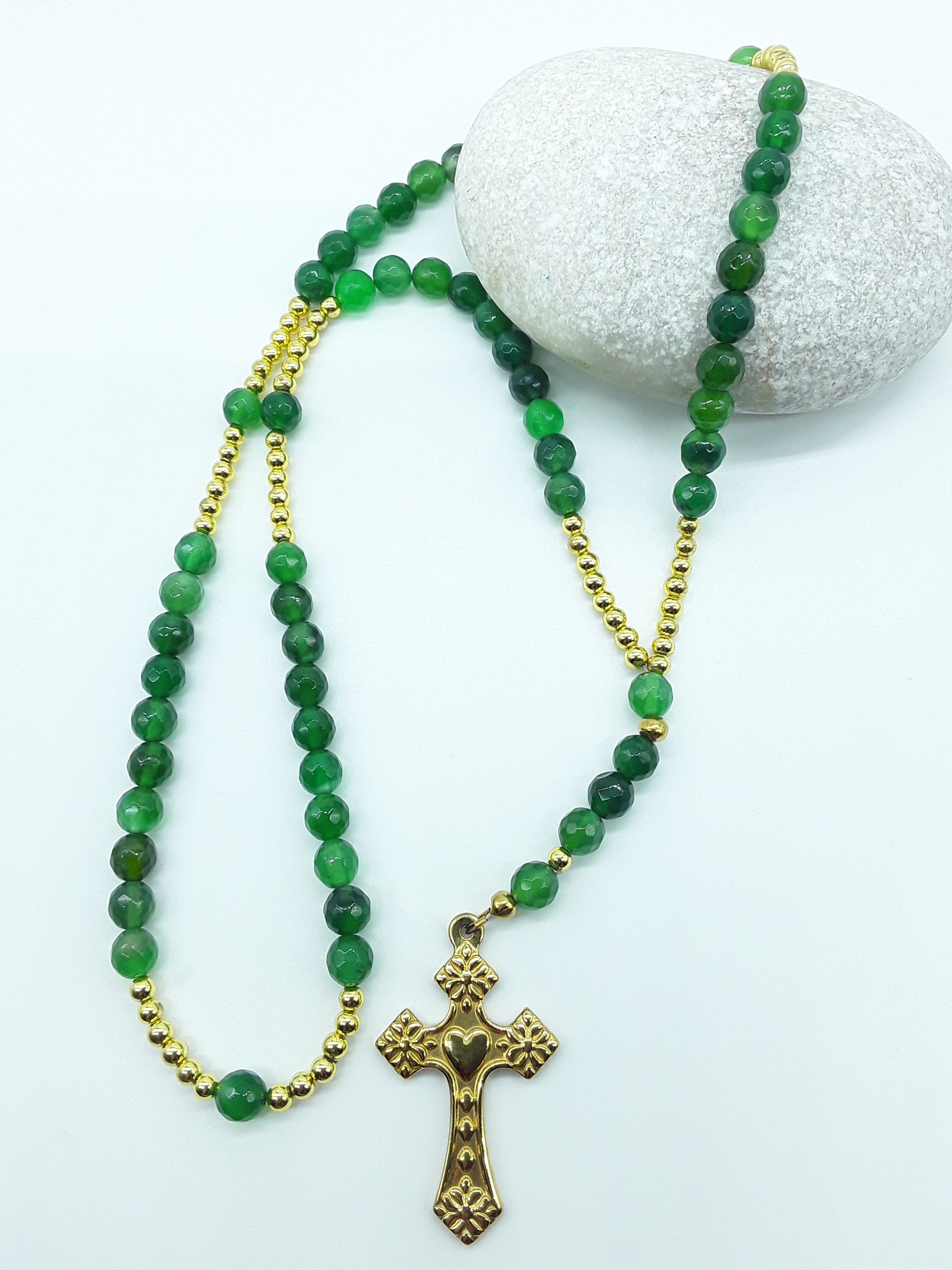 Catholic rosary with natural green agate beads and a stainless steel heart-shaped cross arranged beside a natural stone on a light background.
