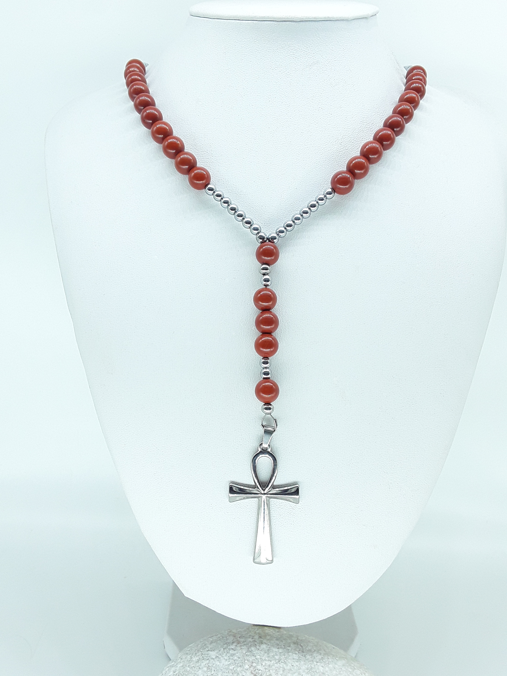 Traditional Catholic rosary made with red jasper beads, hematite spacers, and a stainless steel Ankh cross, displayed on a white bust.
