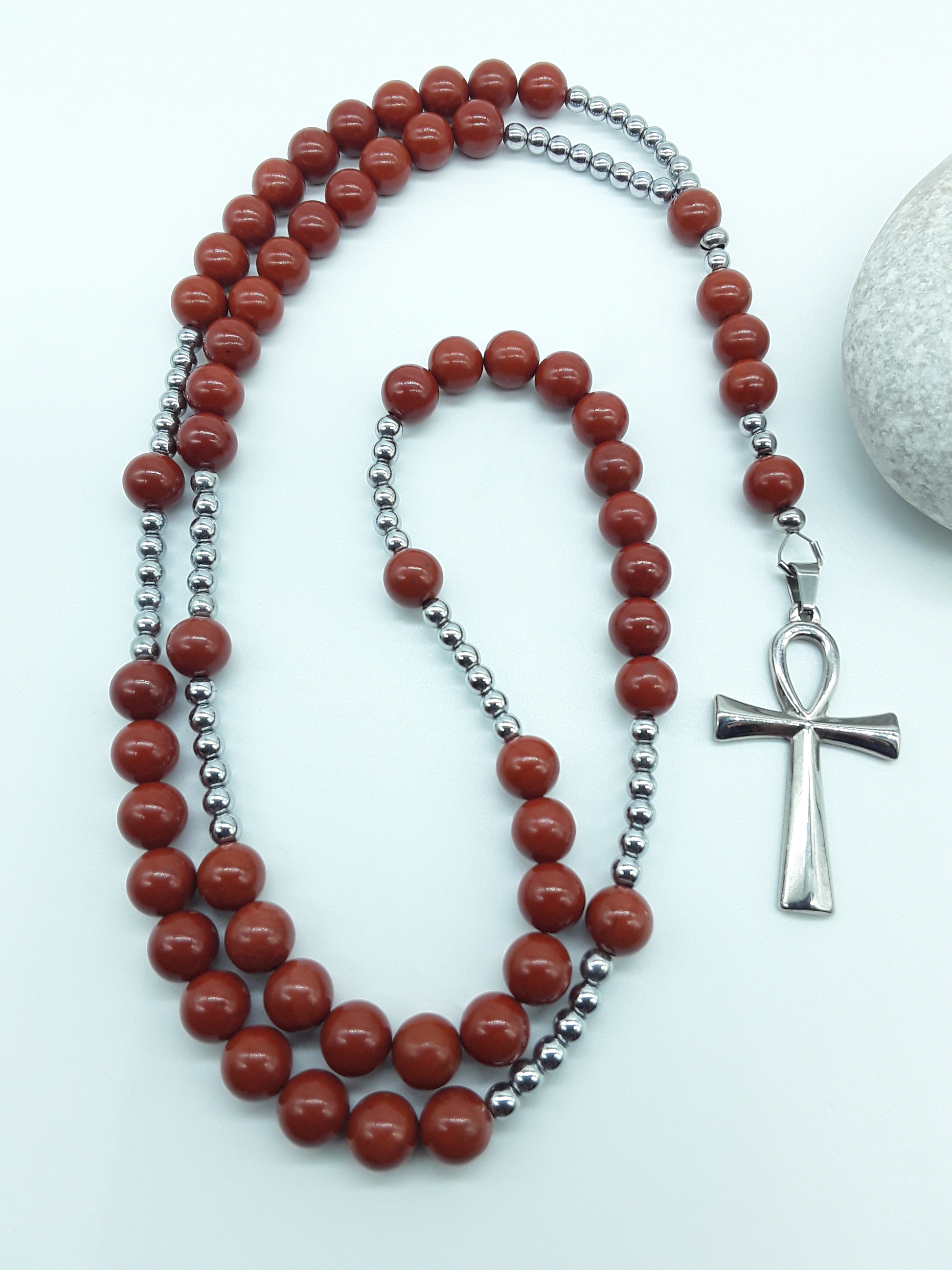 Catholic rosary with red jasper beads, hematite elements, and a stainless steel Ankh cross shown in full length on a light background.
