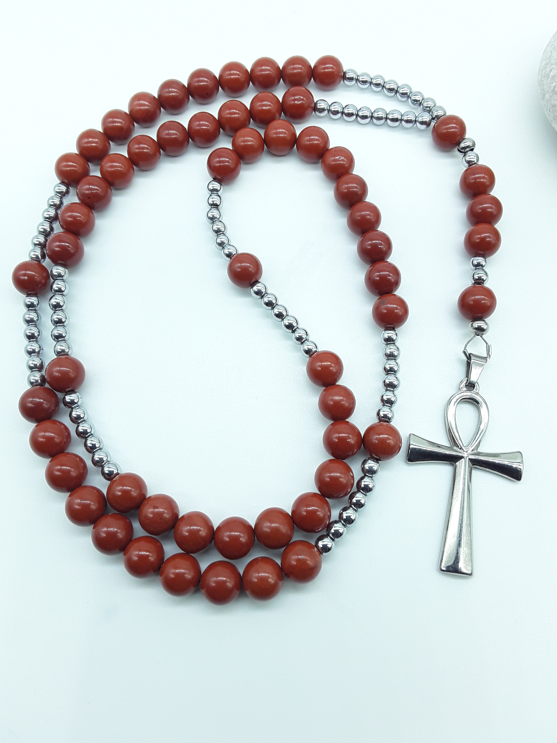 Catholic rosary with red jasper beads, hematite elements, and a stainless steel Ankh cross shown in full length on a light background.
