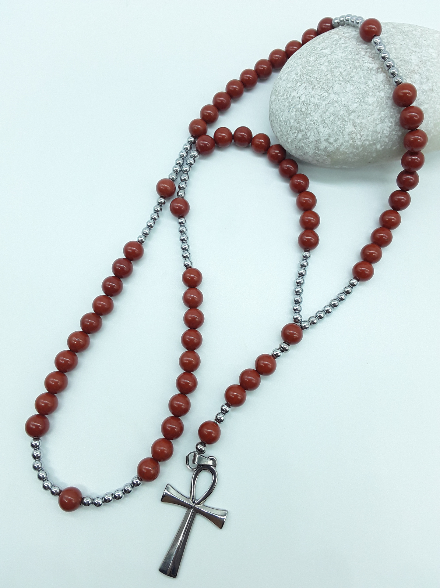 Detail view of a stainless steel Ankh cross attached to a traditional Catholic rosary with red jasper beads.
