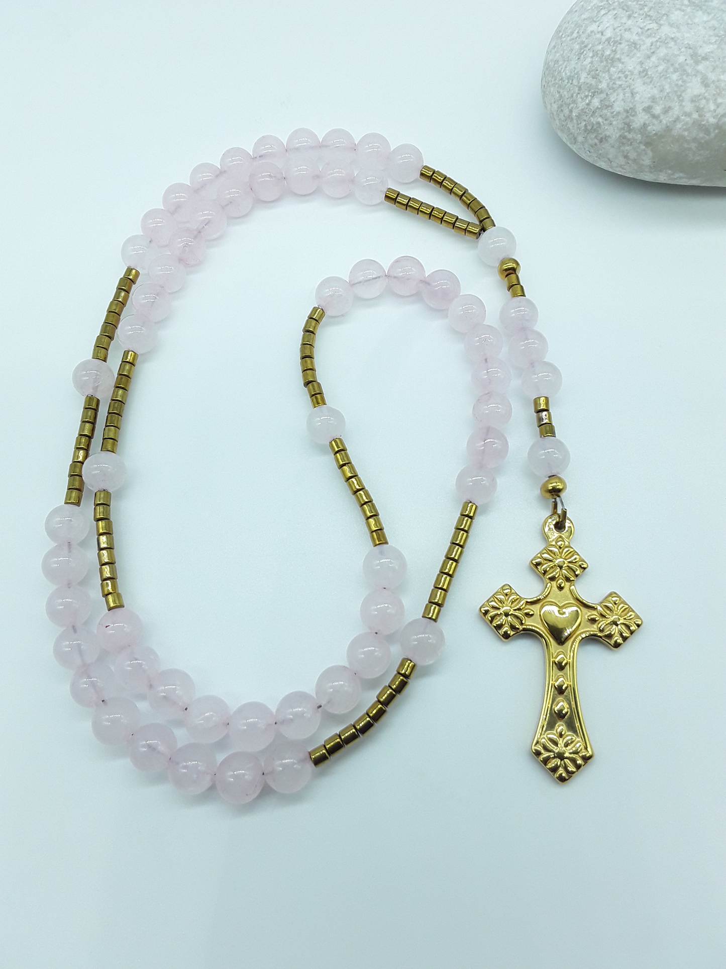 Catholic rosary with rose quartz beads and a stainless steel heart-shaped cross arranged beside a natural stone on a light background.