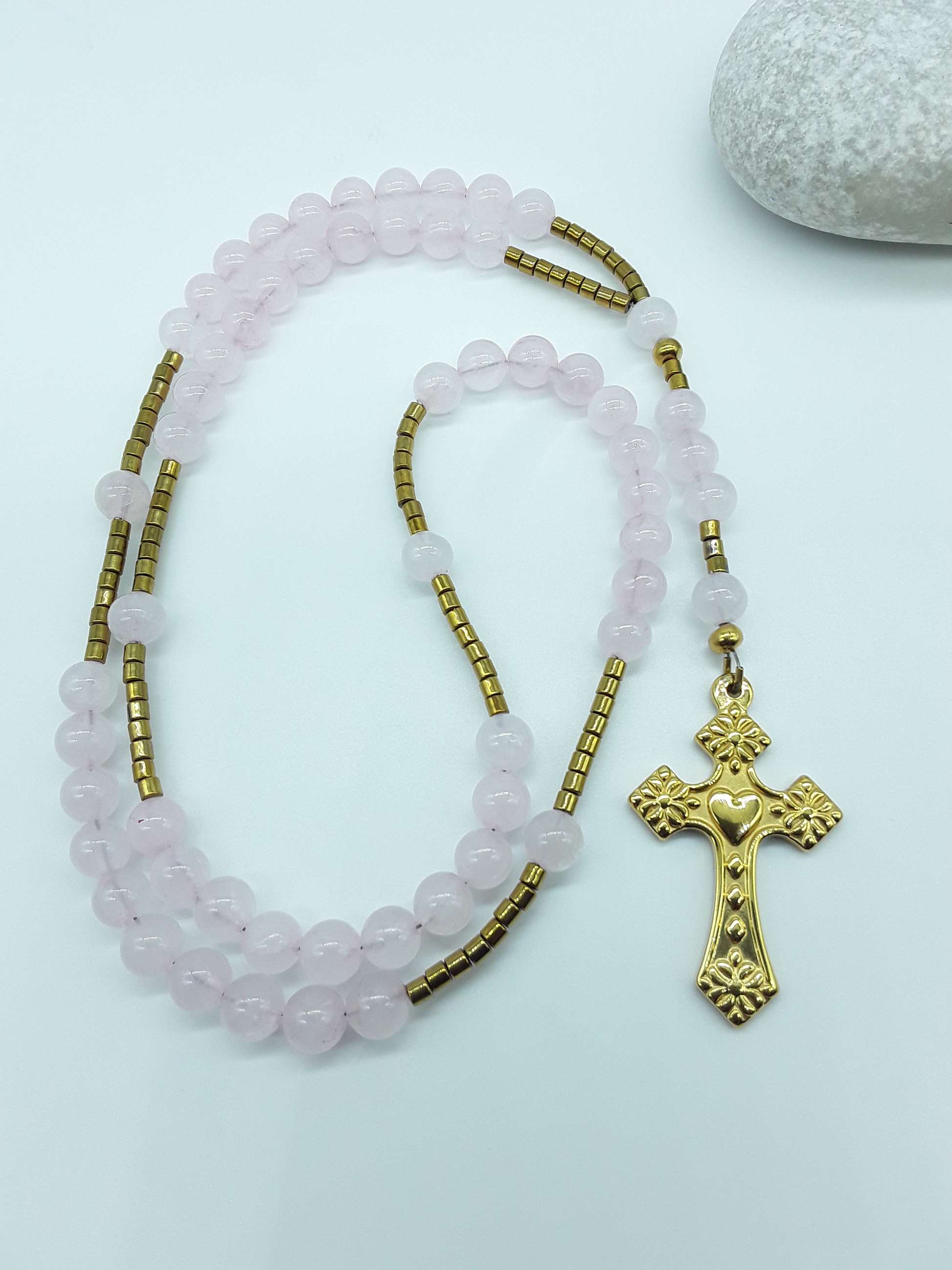 Catholic rosary with rose quartz beads and a stainless steel heart-shaped cross arranged beside a natural stone on a light background.