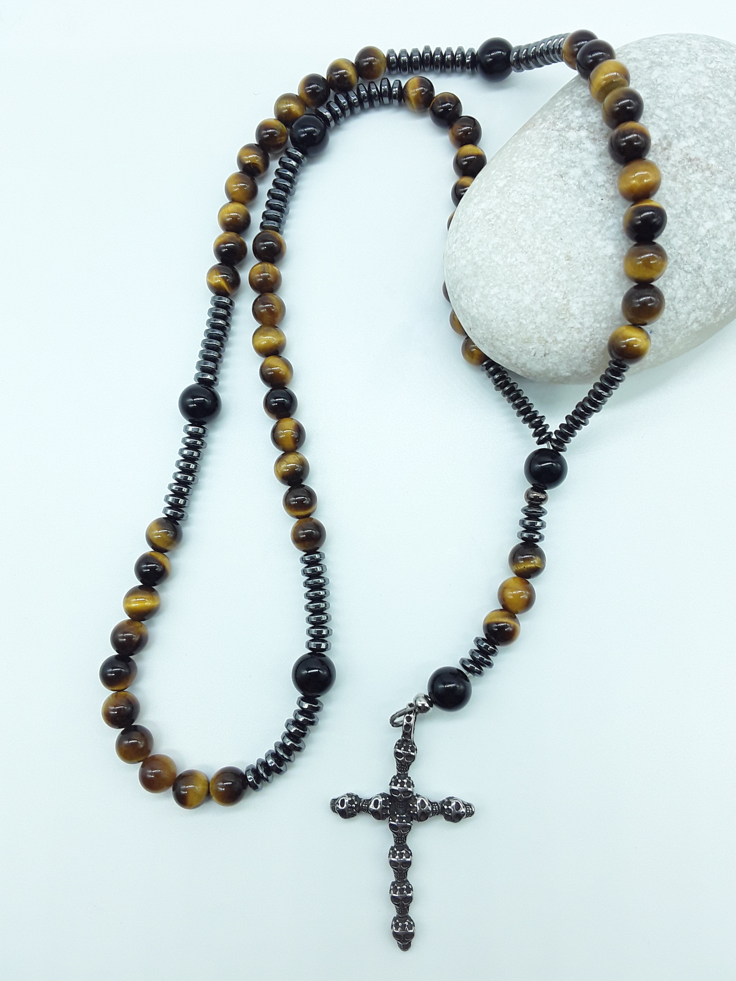 Catholic rosary with tiger’s eye beads, black onyx elements, and a stainless steel skull cross shown in full length on a light background.

