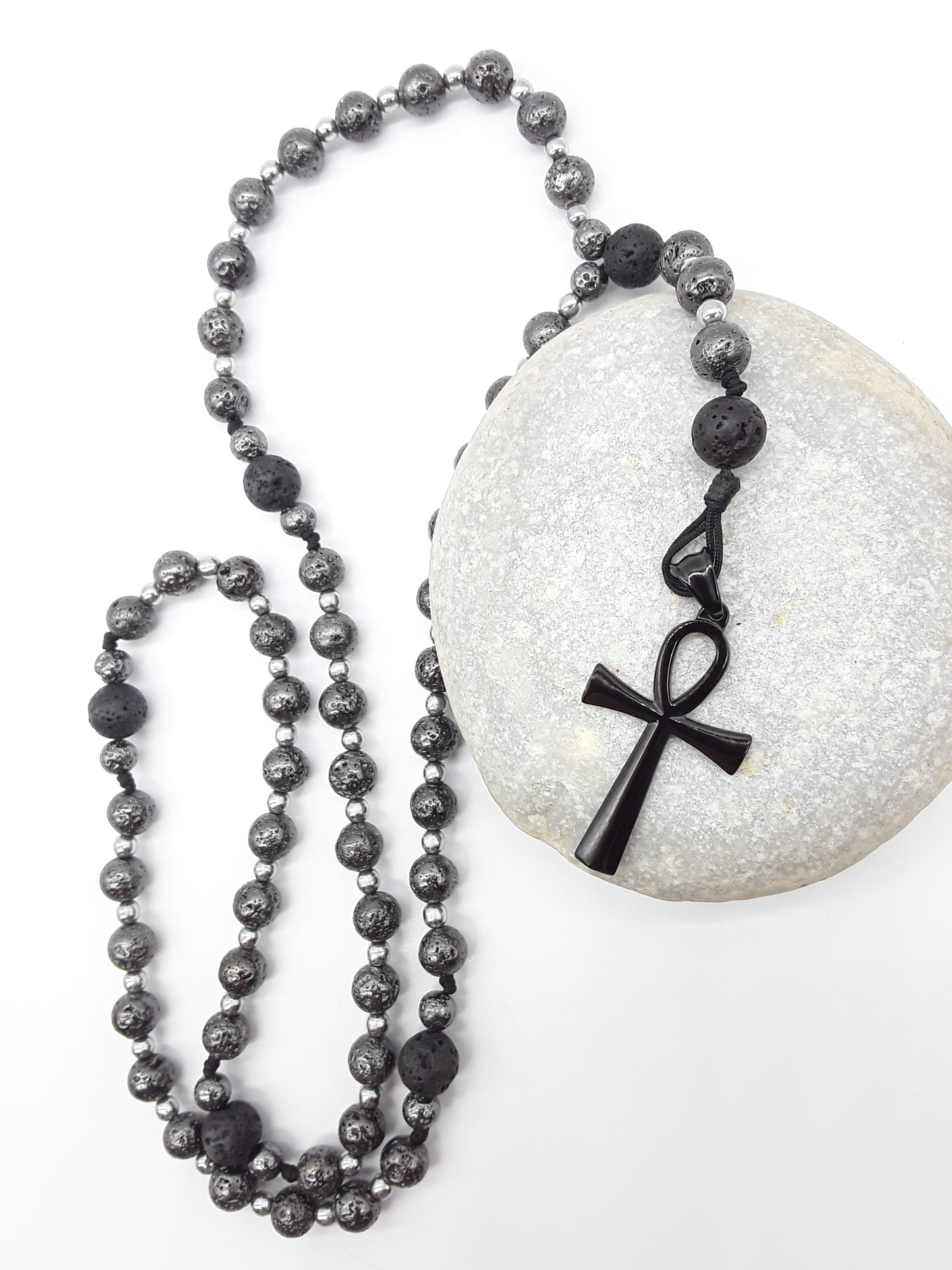 Forged in Continuity lava and hematite Catholic rosary with Ankh cross resting on stone by Polmadeva