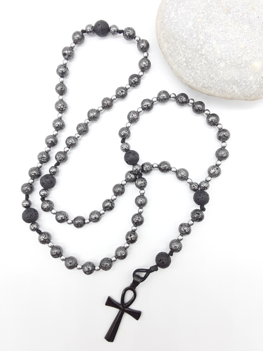 Forged in Continuity lava and hematite Catholic rosary with Ankh cross placed beside stone by Polmadeva