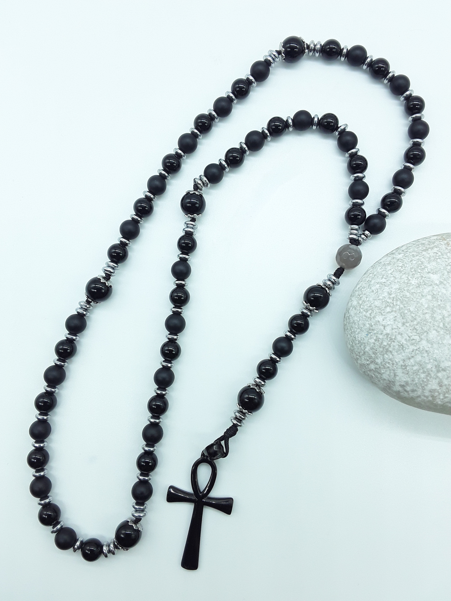 Onyx and Hematite Ankh Catholic Rosary (5 Decade) by Polmadeva placed beside natural stone with black cross.