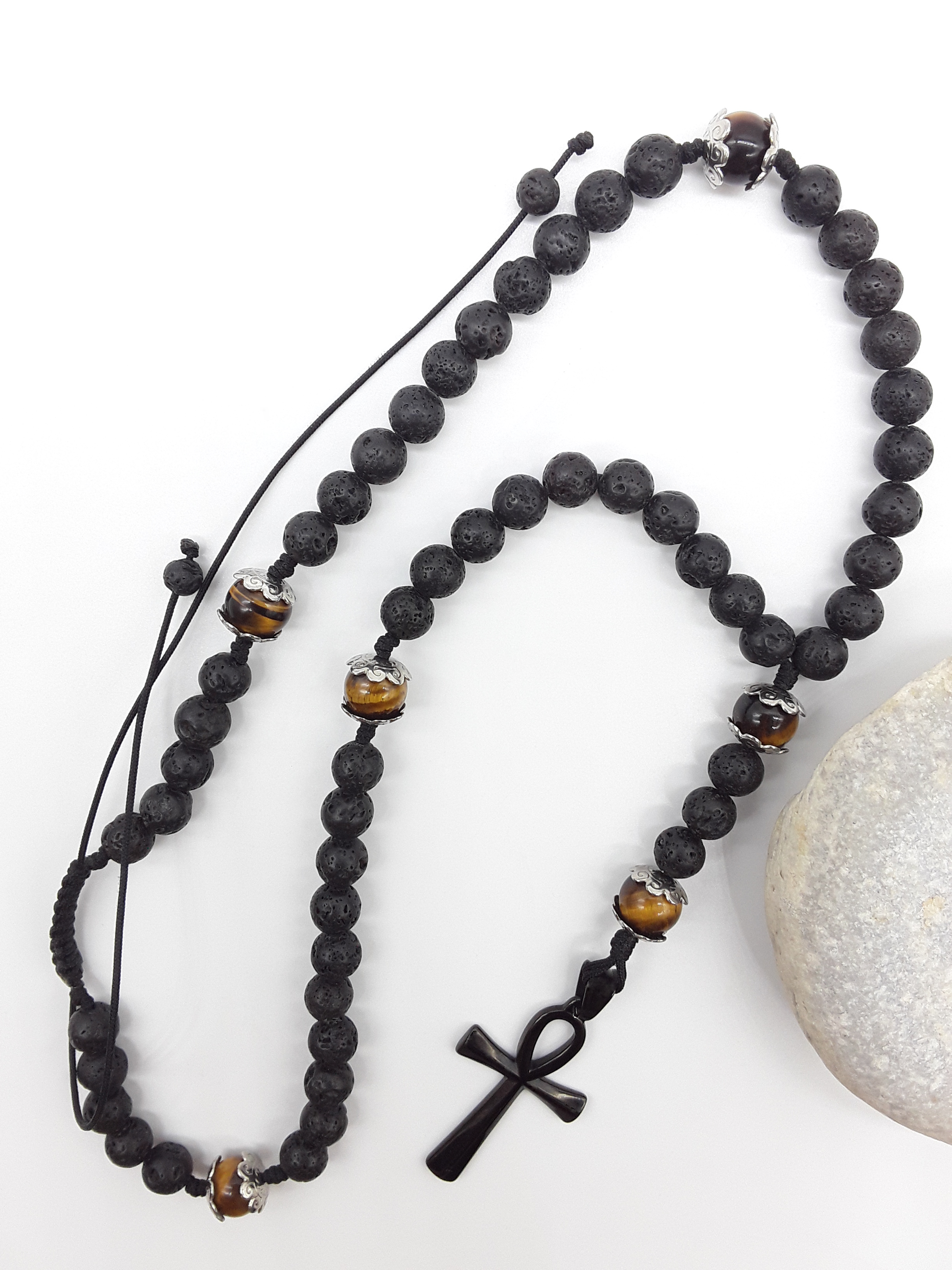 Lava and Tiger’s Eye Ankh Catholic Rosary (5 Decade) by Polmadeva placed beside natural stone with black cross.