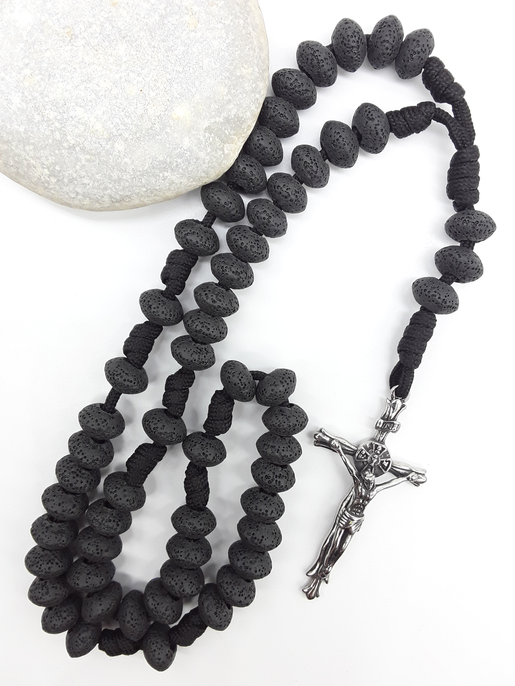 Lava Stone Paracord Catholic Rosary with stainless steel crucifix by Polmadeva resting on natural stone surface.