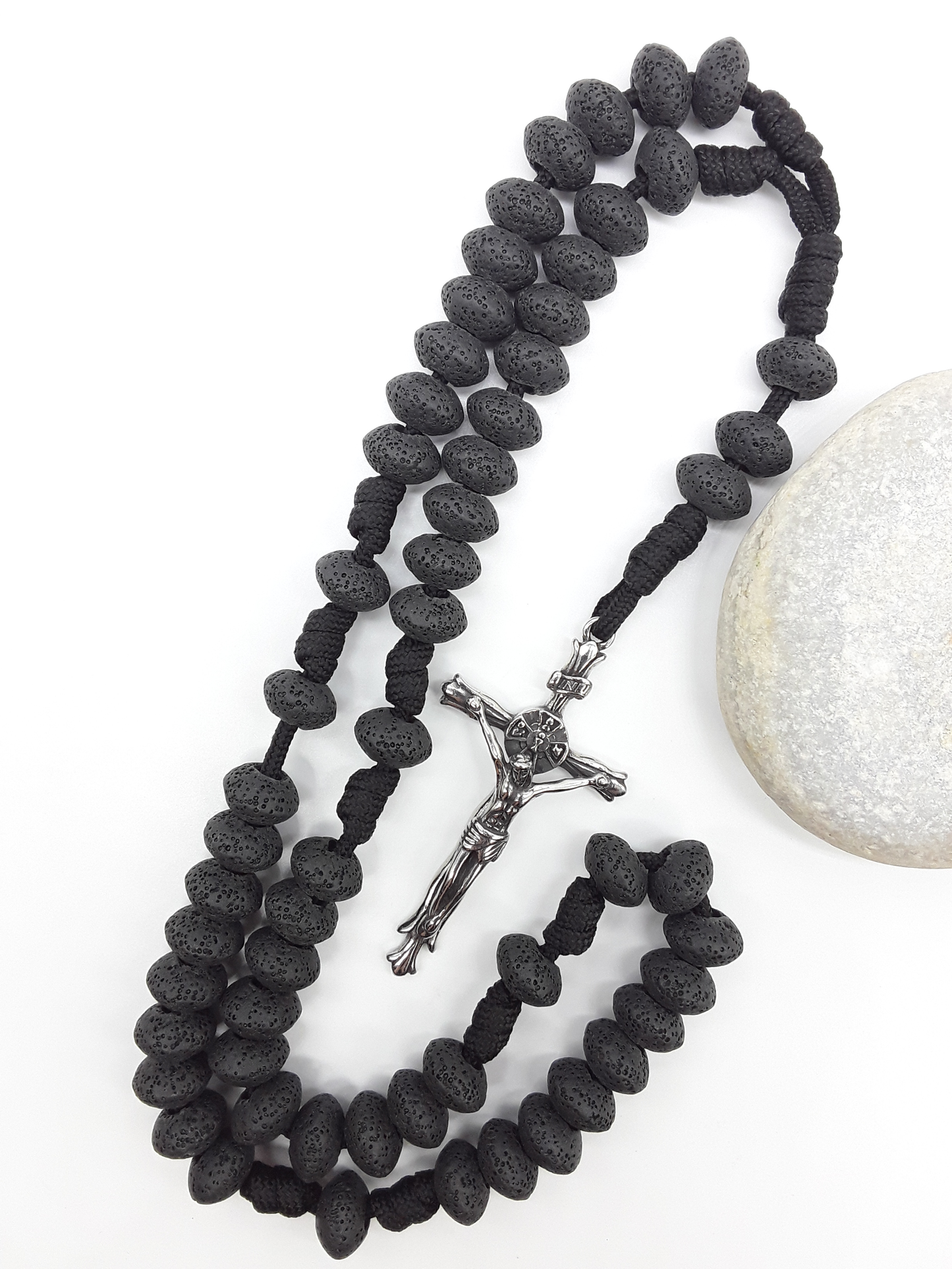Lava Stone Paracord Catholic Rosary with stainless steel crucifix by Polmadeva resting on natural stone surface.