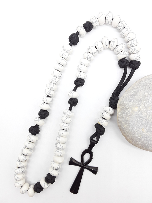 Howlite Paracord Catholic Rosary with Ankh cross by Polmadeva placed beside natural stone, 5-decade design.