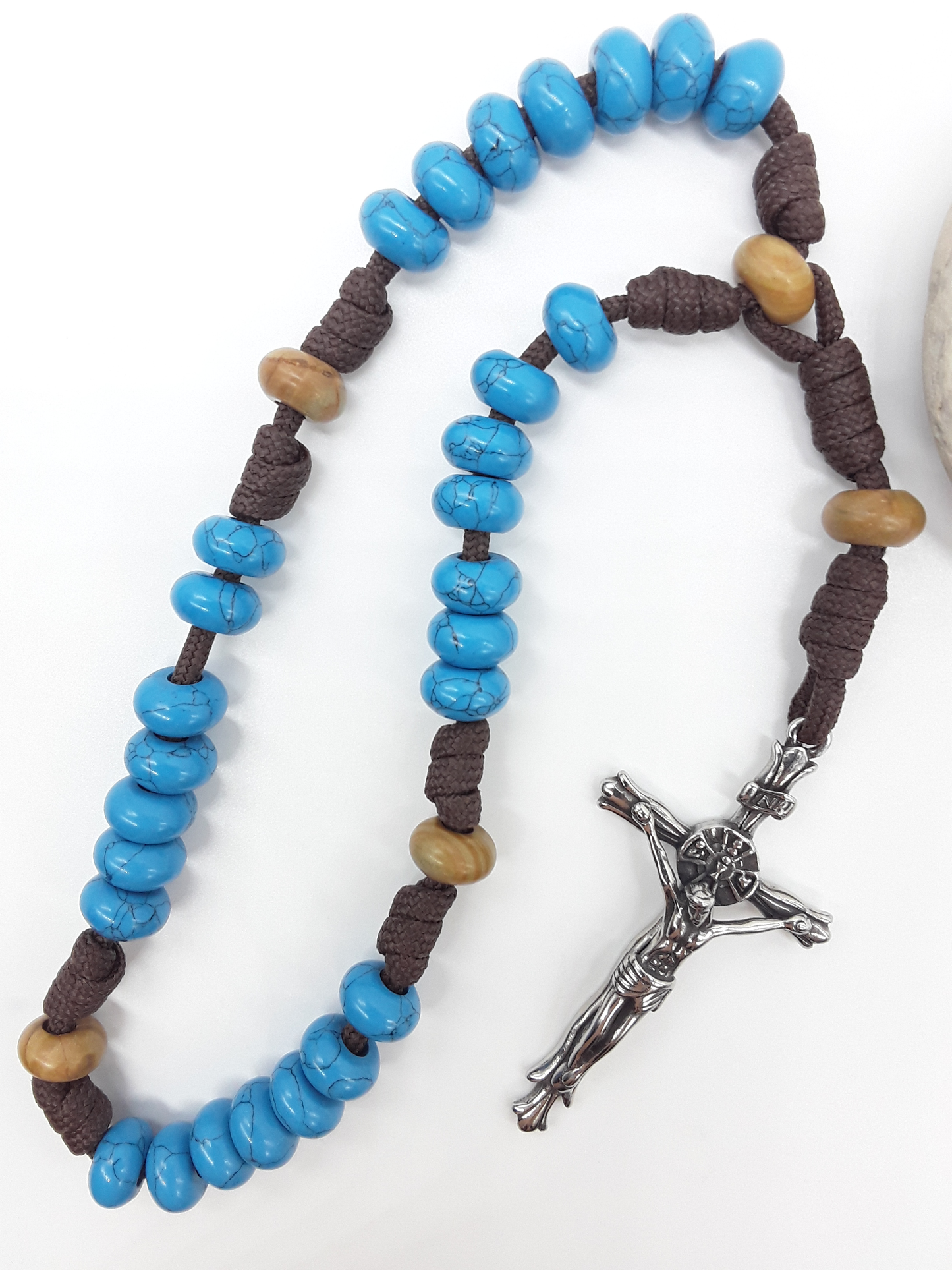 The Anchored Way Paracord Anglican Prayer Beads by Polmadeva beside natural stone, featuring Turquoise, Wood Lace beads, and crucifix