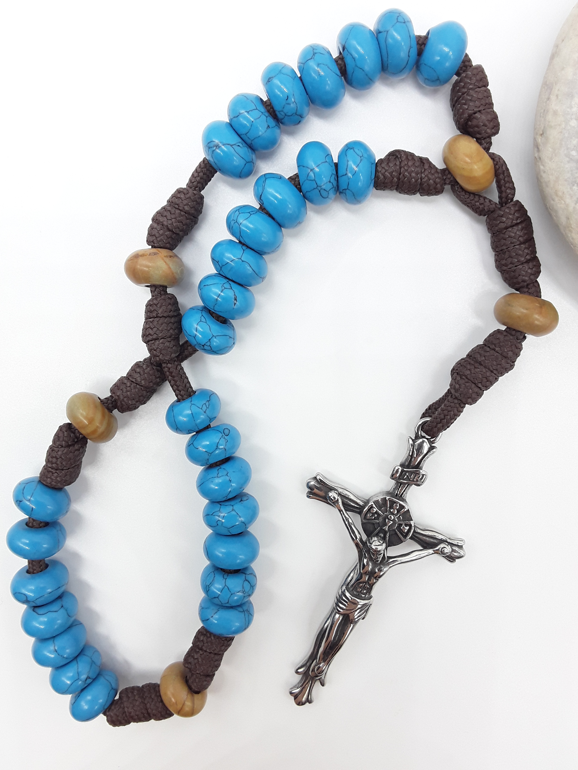 The Anchored Way Paracord Anglican Prayer Beads by Polmadeva beside natural stone, featuring Turquoise, Wood Lace beads, and crucifix