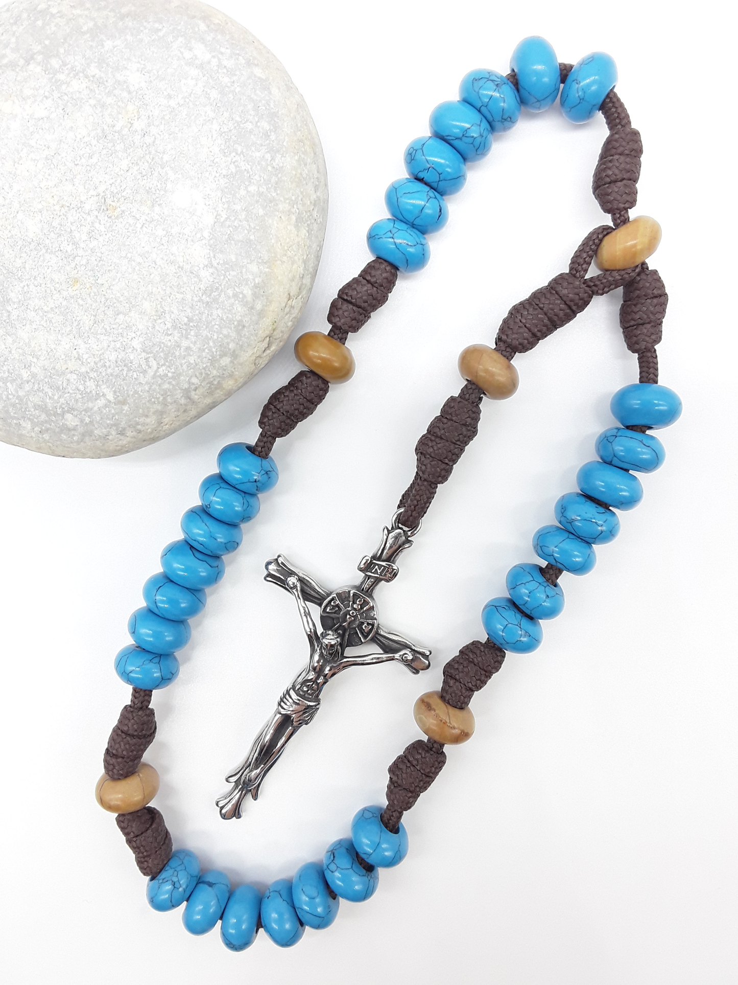The Anchored Way Paracord Anglican Prayer Beads by Polmadeva beside natural stone, featuring Turquoise, Wood Lace beads, and crucifix