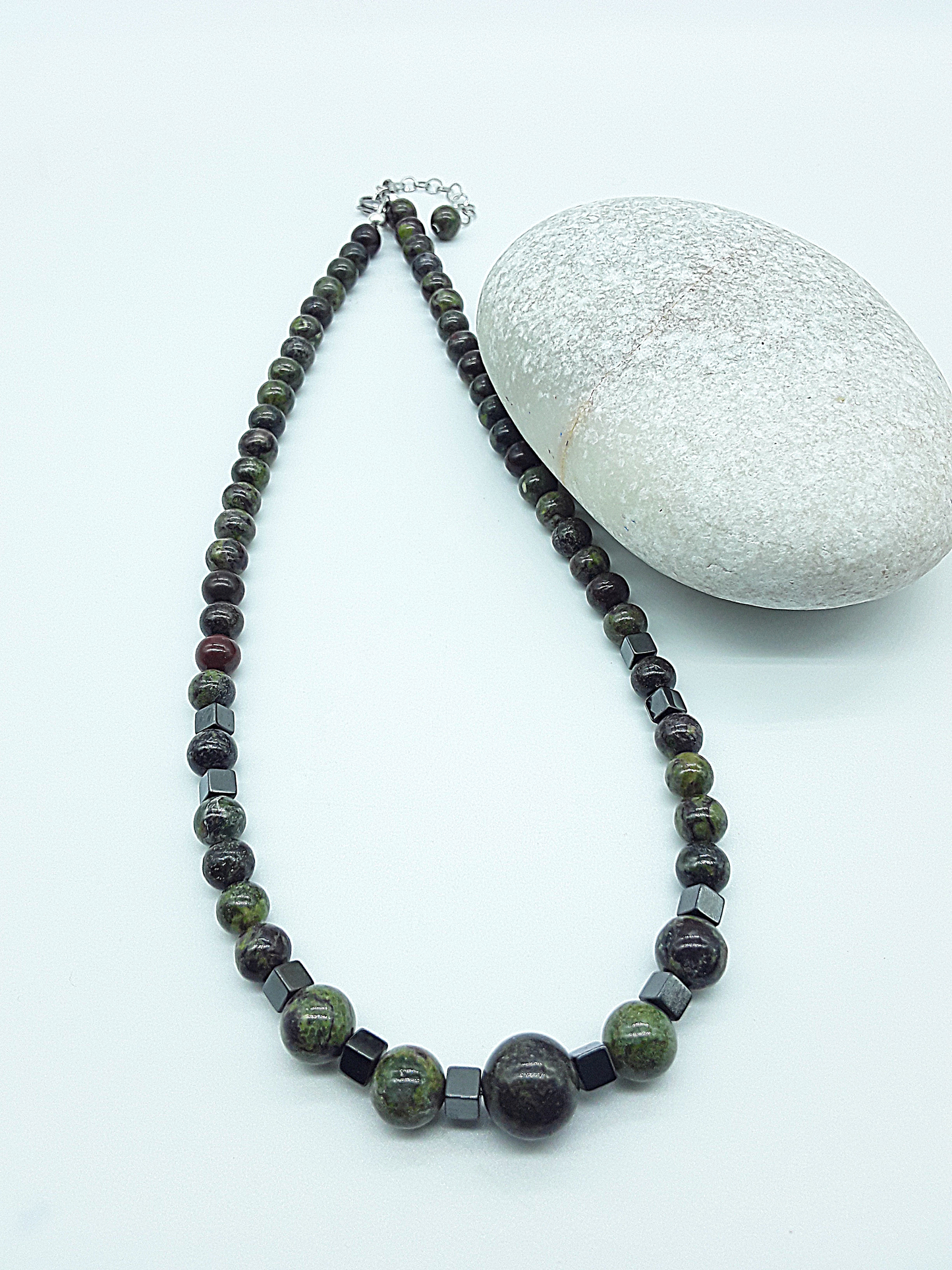 The Emberwake Necklace by Polmadeva arranged beside a natural stone on a light background, handcrafted with Dragon Blood Stone beads and hematite accents, a Courage Rising talisman representing awakened inner fire, grounded strength, and resilience.