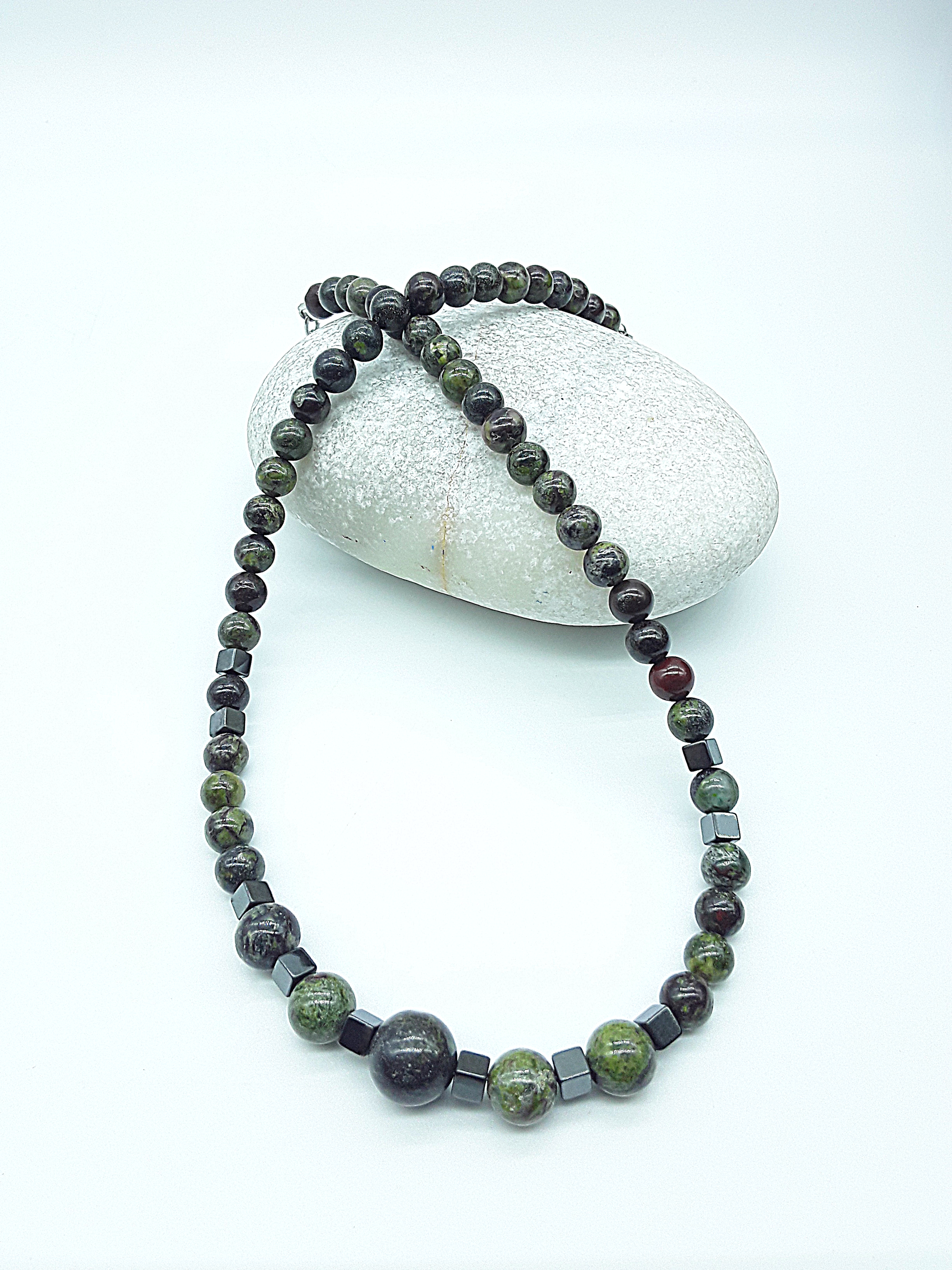 The Emberwake Necklace by Polmadeva draped over a natural stone on a light background, handcrafted with Dragon Blood Stone beads and hematite accents, a Courage Rising talisman symbolizing awakened inner fire, grounded power, and resilient strength.