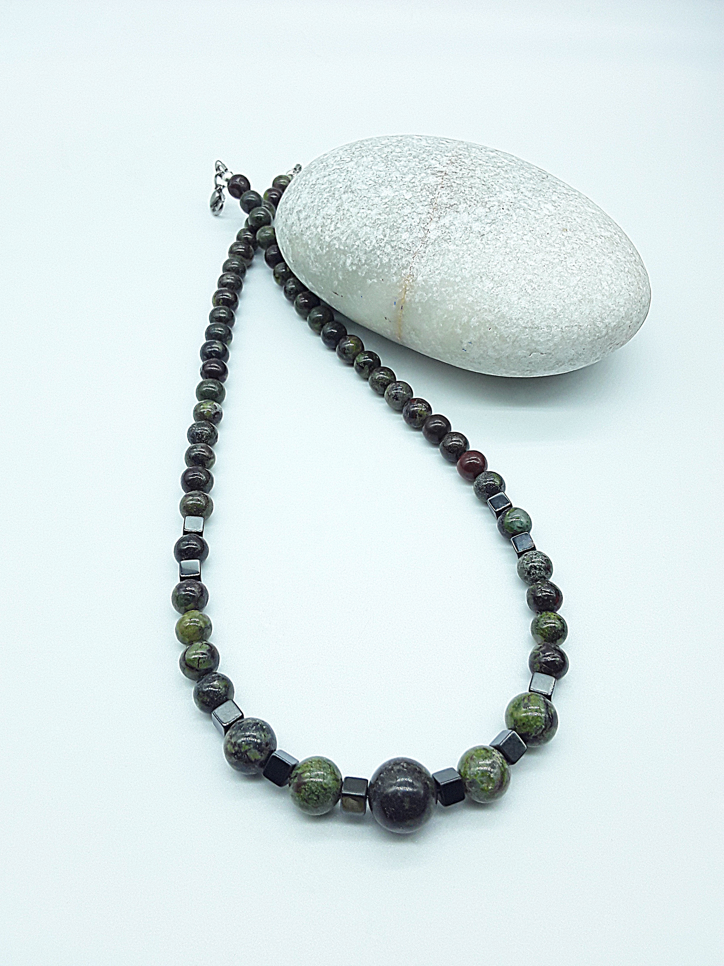 The Emberwake Necklace by Polmadeva resting beside a natural stone on a light background, handcrafted with Dragon Blood Stone beads and hematite accents, a Courage Rising talisman symbolizing awakened inner fire, grounded strength, and steady resilience.