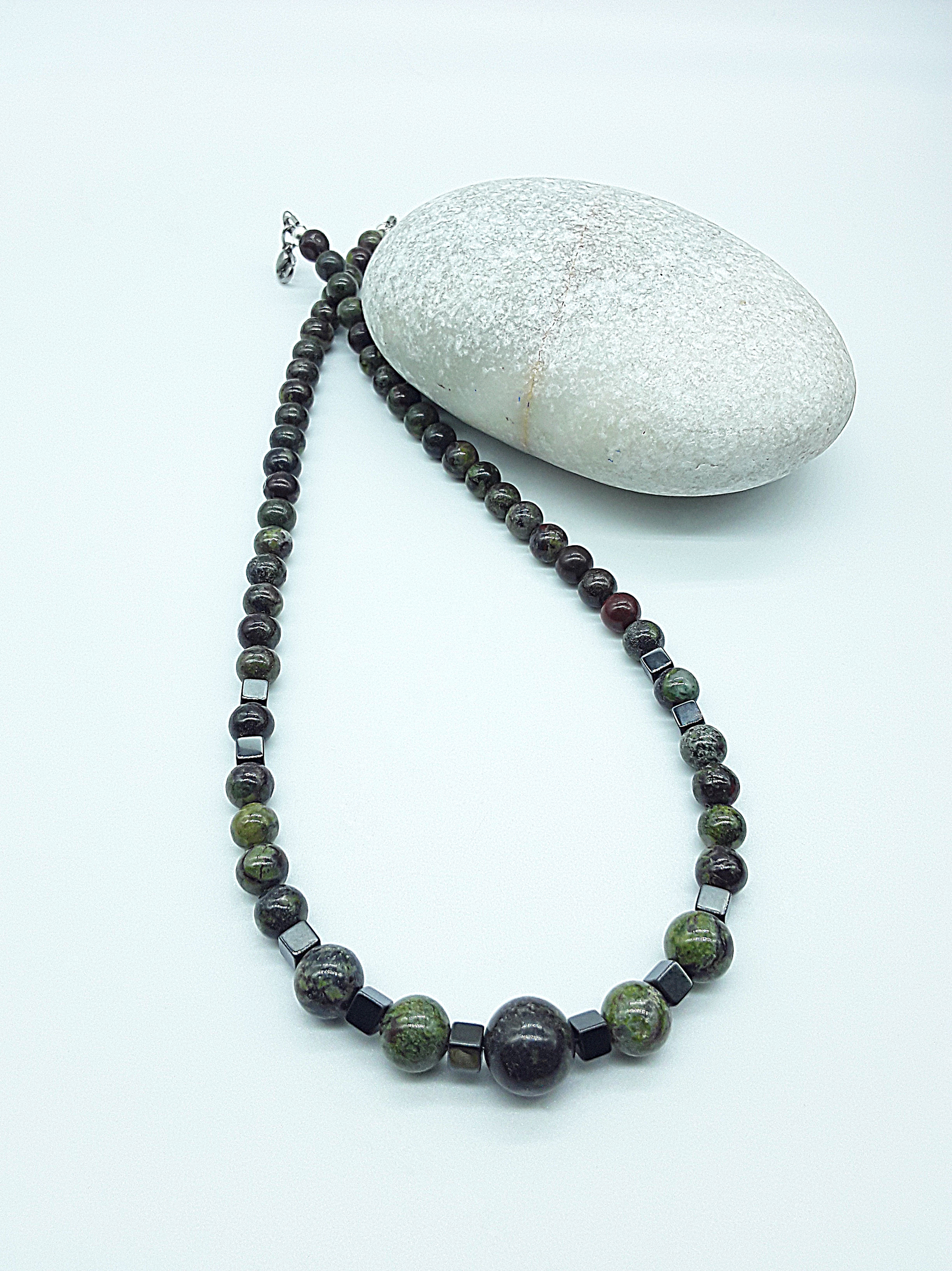 The Emberwake Necklace by Polmadeva resting beside a natural stone on a light background, handcrafted with Dragon Blood Stone beads and hematite accents, a Courage Rising talisman symbolizing awakened inner fire, grounded strength, and steady resilience.