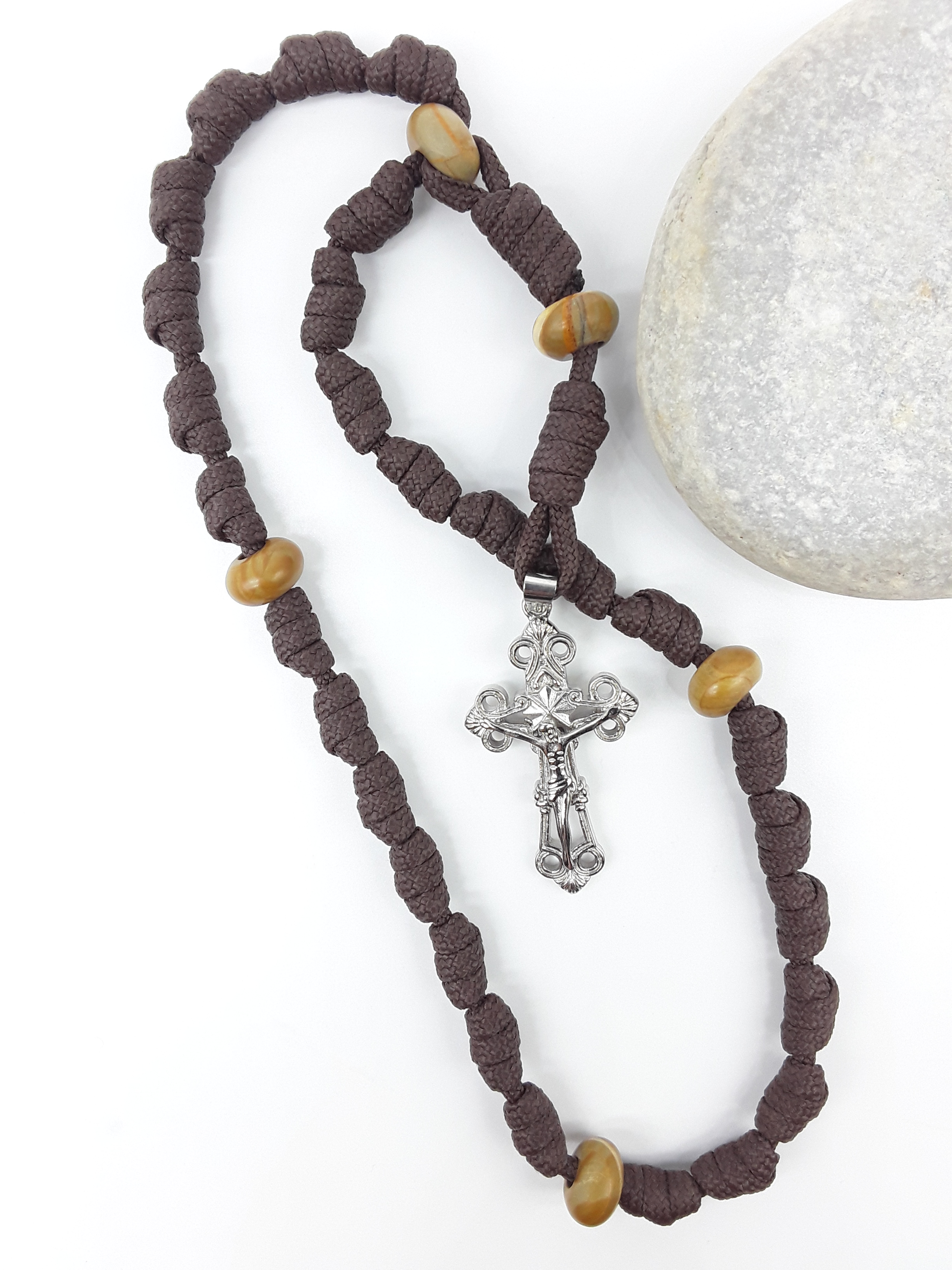 The Enduring Light Anglican Rosary by Polmadeva with hand-knotted brown paracord, Natural Wood elements, and Eternal Circle Crucifix beside stone.