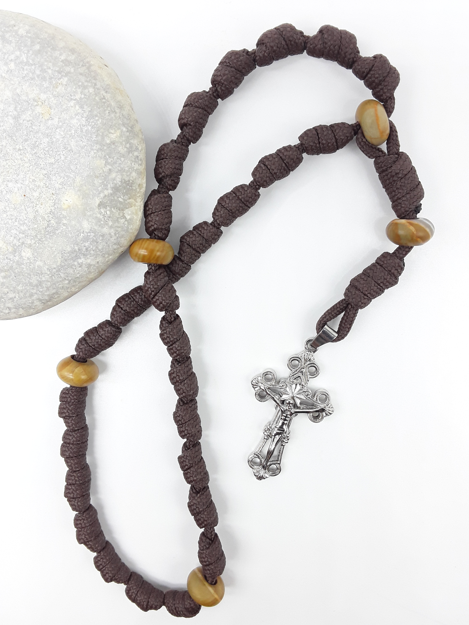 The Enduring Light Anglican Rosary by Polmadeva with hand-knotted brown paracord, Natural Wood elements, and Eternal Circle Crucifix beside stone.