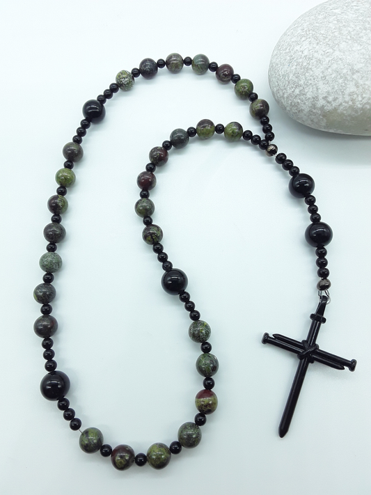 The Enduring Root Anglican Prayer Beads by Polmadeva beside natural stone, featuring Dragon Blood, Black Onyx, and nail-style cross
