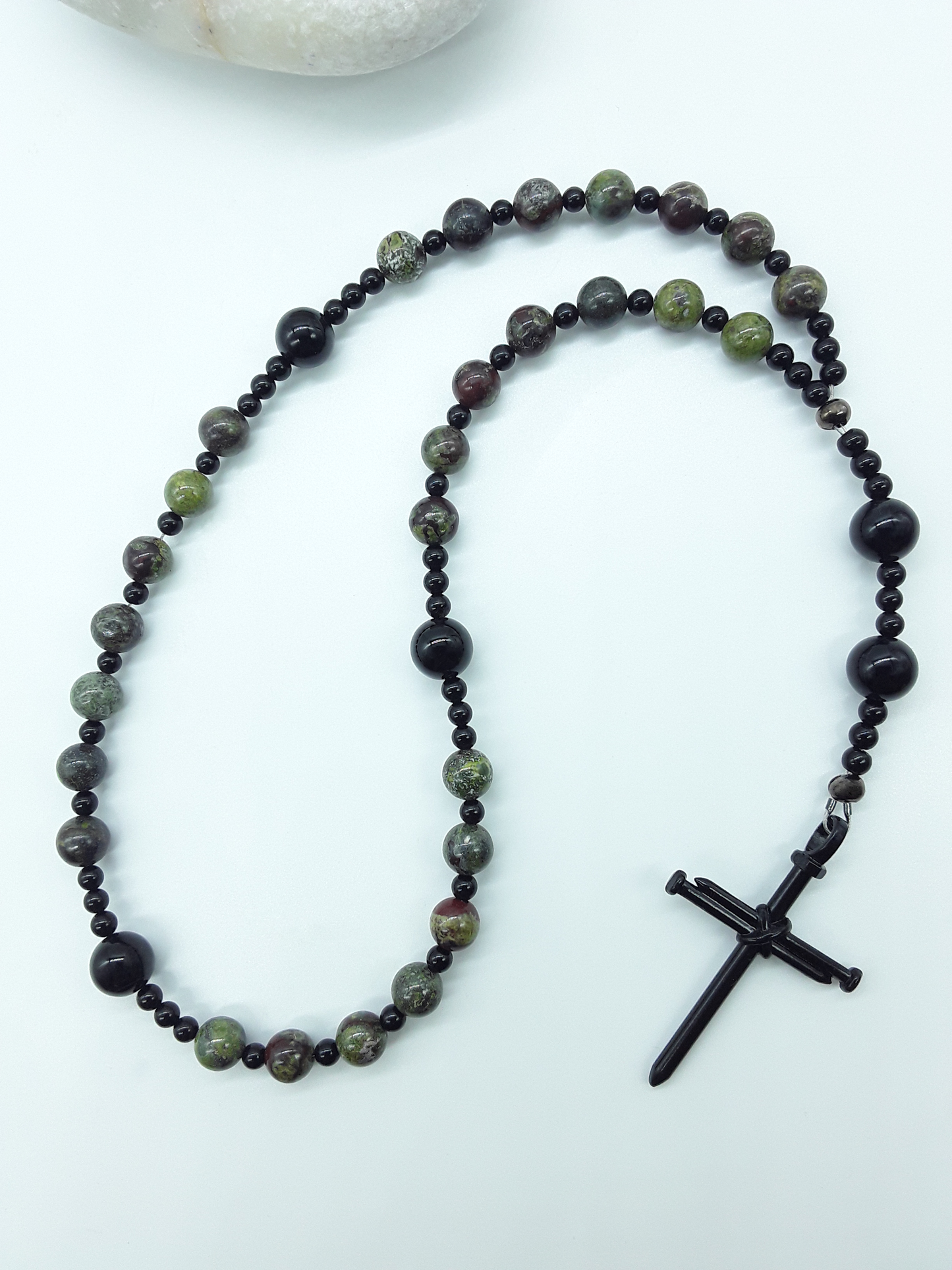 The Enduring Root Anglican Prayer Beads by Polmadeva beside natural stone, featuring Dragon Blood, Black Onyx, and nail-style cross