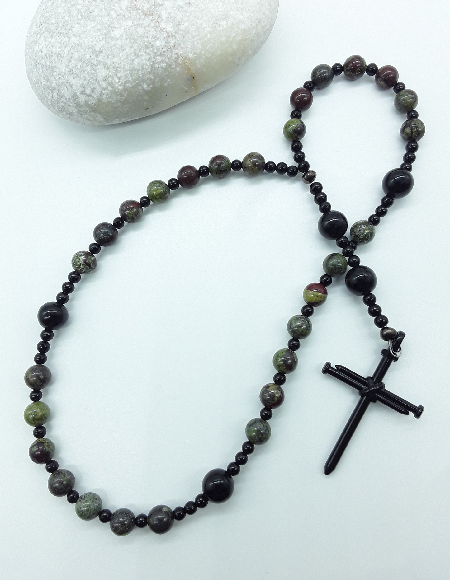 The Enduring Root Anglican Prayer Beads by Polmadeva beside natural stone, featuring Dragon Blood, Black Onyx, and nail-style cross