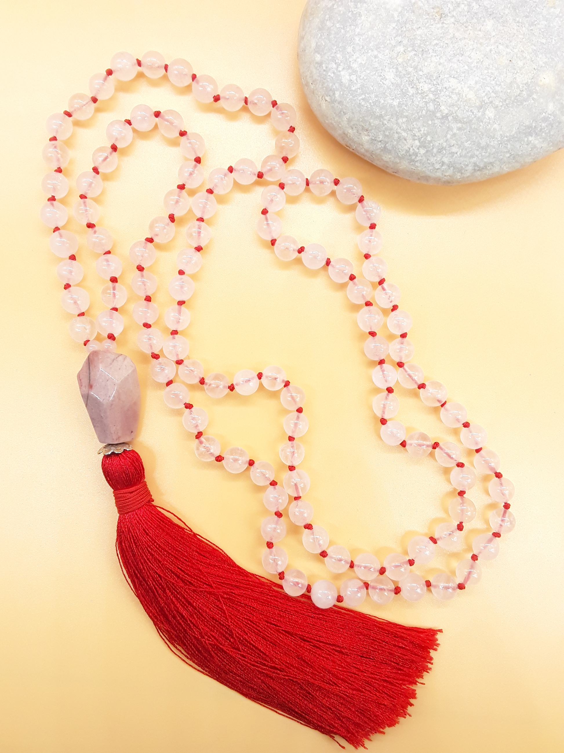 Rose Quartz Mala Beads with Agate guru bead and red tassel placed beside white stone on soft background, Polmadeva