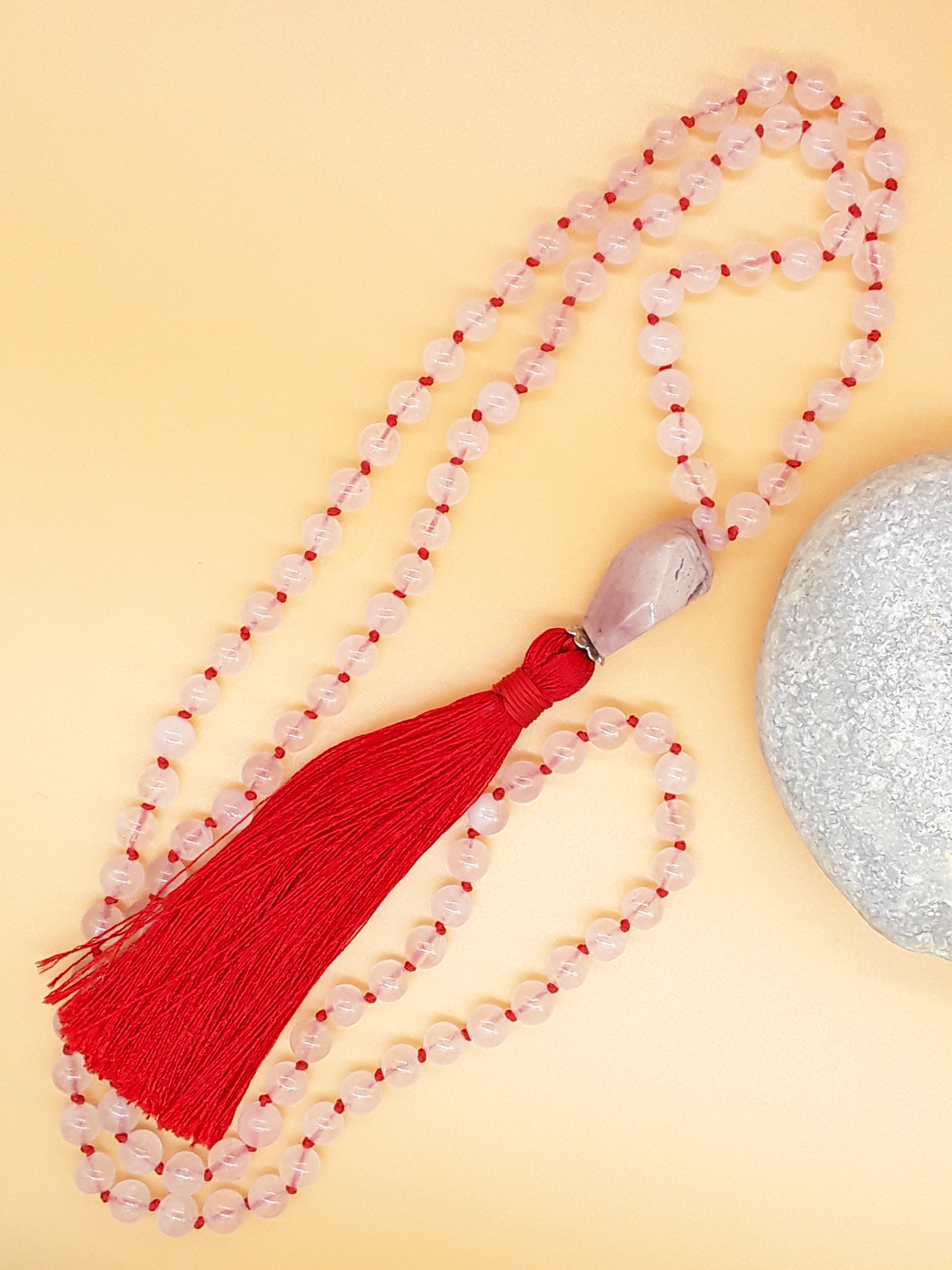 Rose Quartz Mala Beads with Agate guru bead and red tassel placed beside white stone on soft background, Polmadeva
