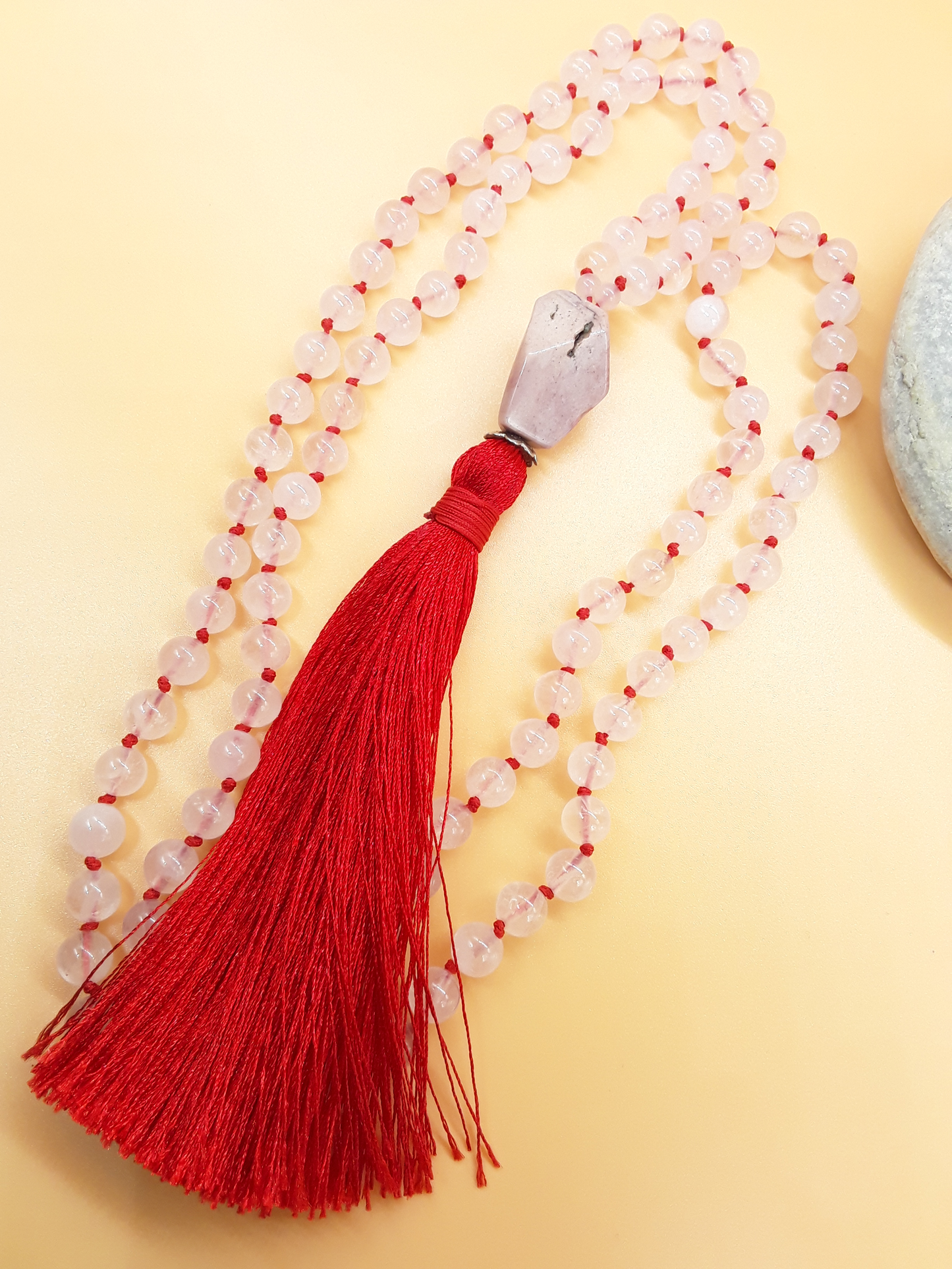 Rose Quartz Mala Beads with Agate guru bead and red tassel placed beside white stone on soft background, Polmadeva