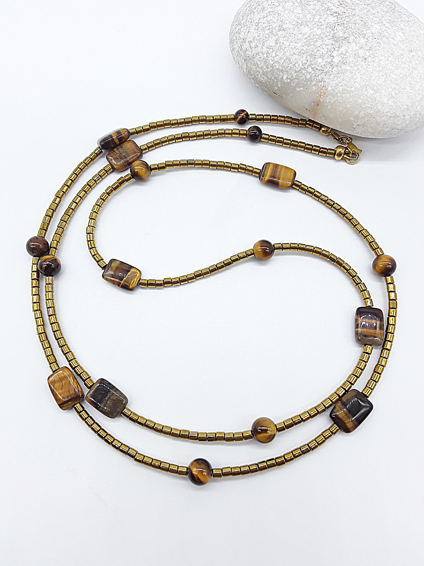 Gold-toned beaded necklace with Tiger’s Eye and hematite beads placed on a white surface next to a round stone.