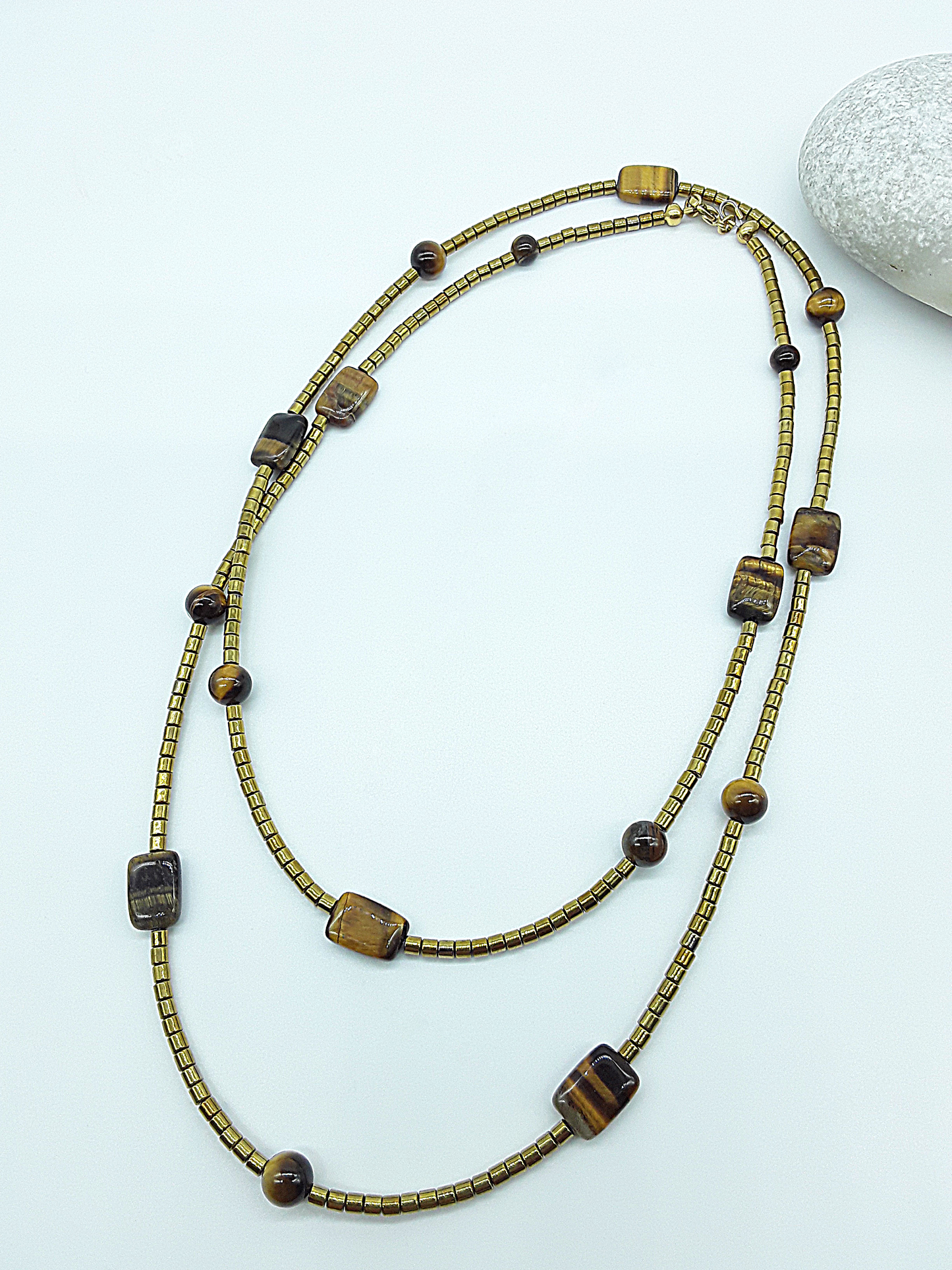 Gold-toned beaded necklace featuring round and rectangular Tiger’s Eye beads displayed on a white background next to a stone.