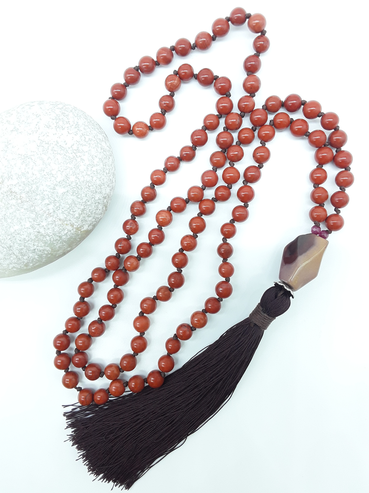 Red Jasper Mala Beads with Agate guru bead and dark tassel placed beside white stone on white background, Polmadeva