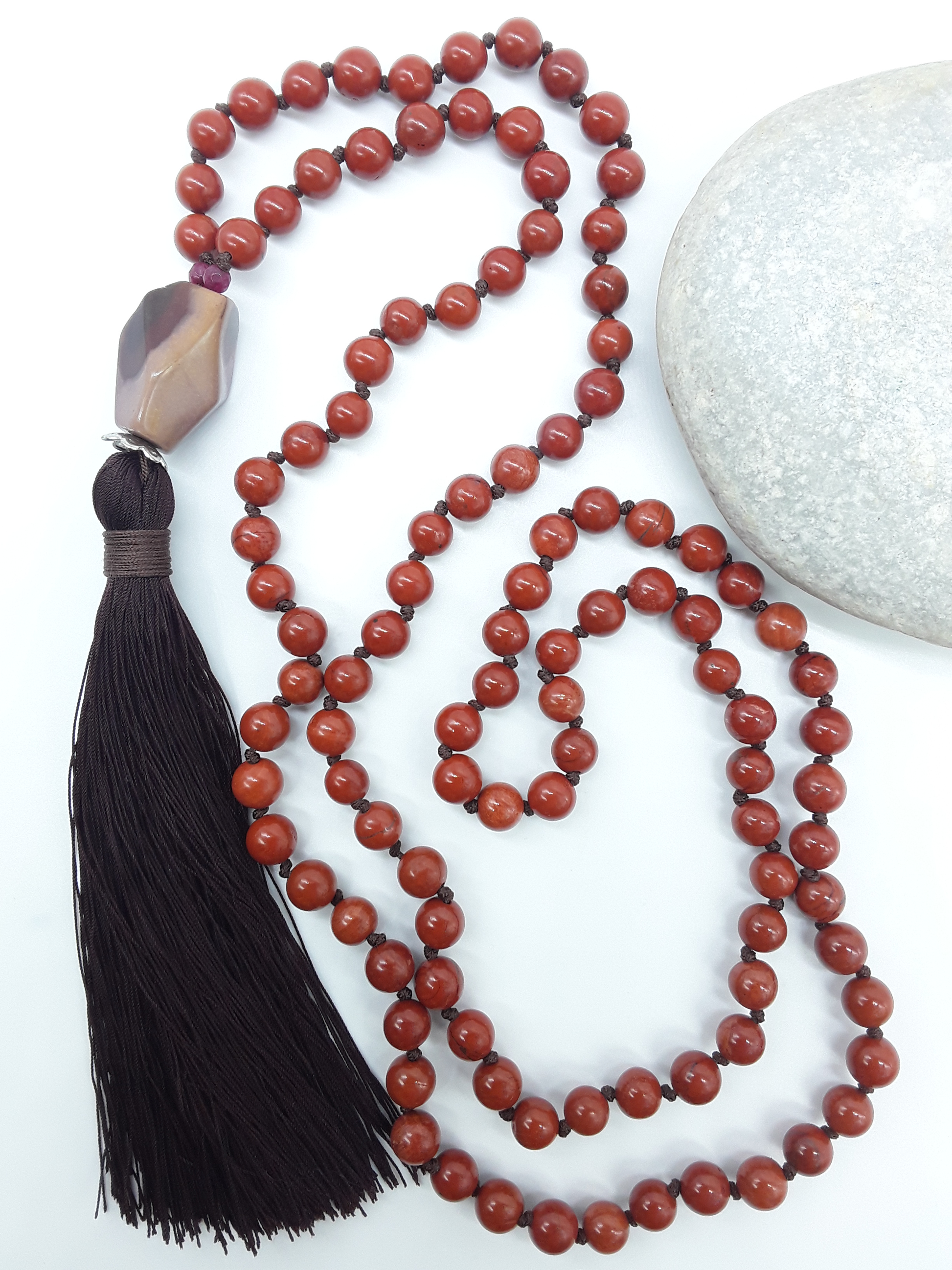 Red Jasper Mala Beads with Agate guru bead and dark tassel placed beside white stone on white background, Polmadeva