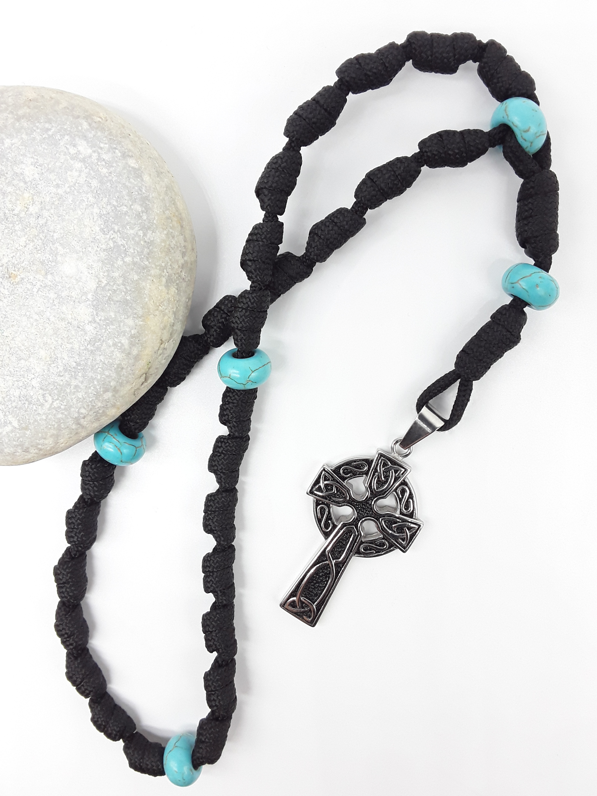 The Guiding Tide Celtic Trinity Anglican Rosary by Polmadeva with hand-knotted black paracord, Turquoise elements, and Celtic Trinity Cross beside stone.